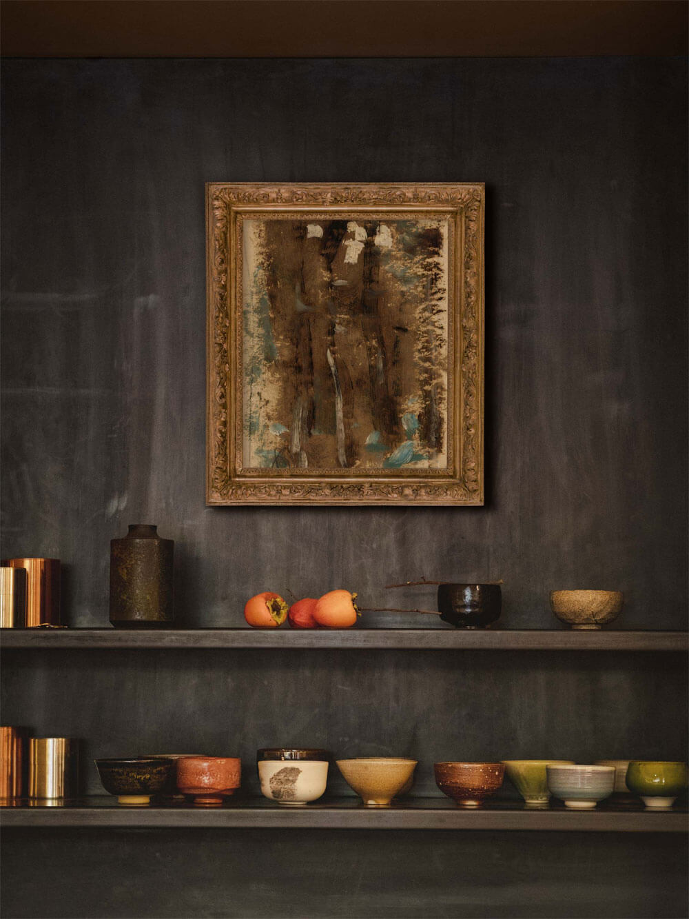 Abstraktes Kunstposter in antikem Rahmen auf einem dunkelgrauen Regal mit handgefertigten Schalen und Persimonen als Dekoration.
