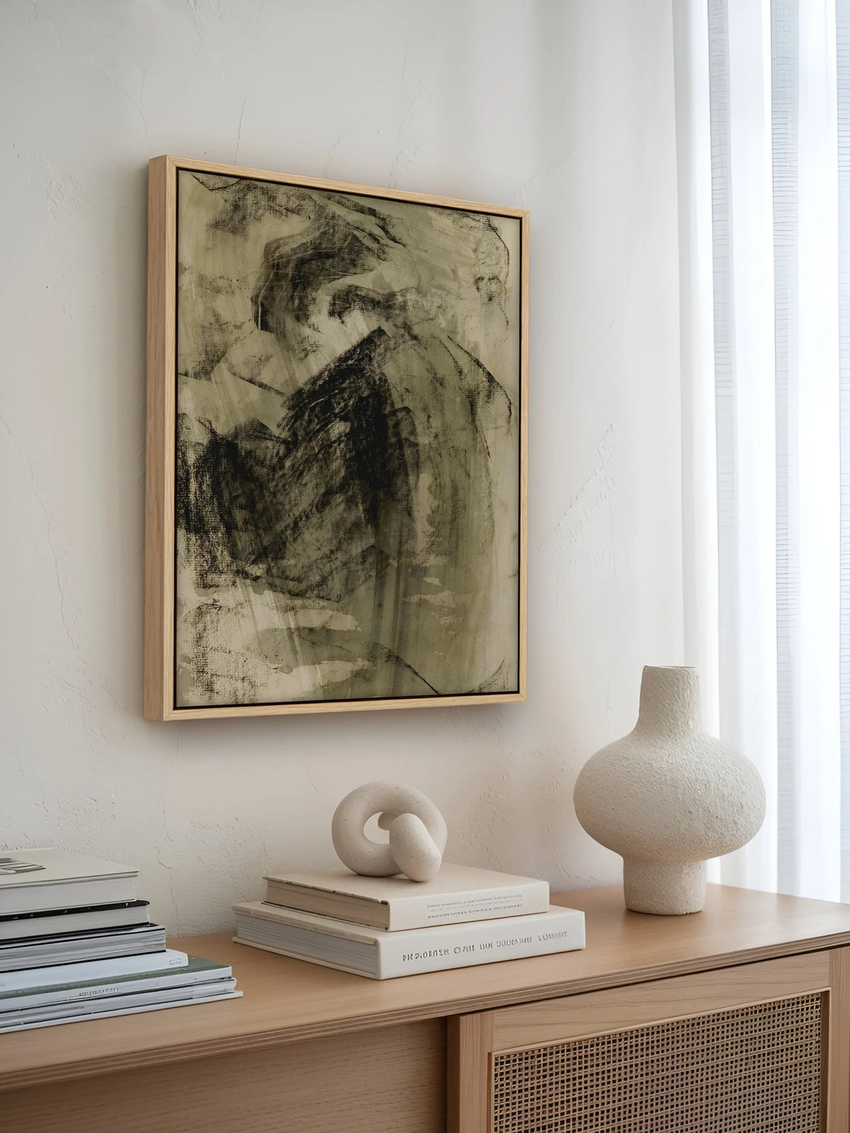 Abstraktes Leinwandbild im Naturholzrahmen über einem Sideboard mit Rattanfront, Büchern und Keramikvase. Das Wabi Sabi Wandbild passt mit seinen Oliv- und Schwarztönen zur hellen Japandi Einrichtung.