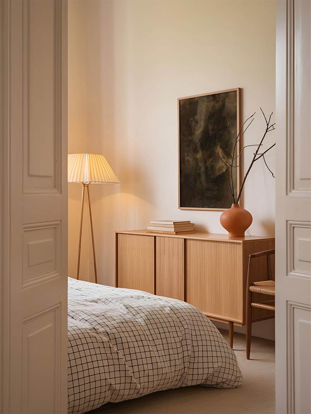 Abstrakter Kunstdruck in dunklen Braun- und Olivtönen hängt über einem hellen Sideboard im ruhigen Schlafzimmer mit Holz, Leinenbettwäsche und warmer Beleuchtung. Das Wandbild ergänzt eine minimalistische, wabi-sabi-inspirierte Einrichtung mit natürlicher Ruhe.