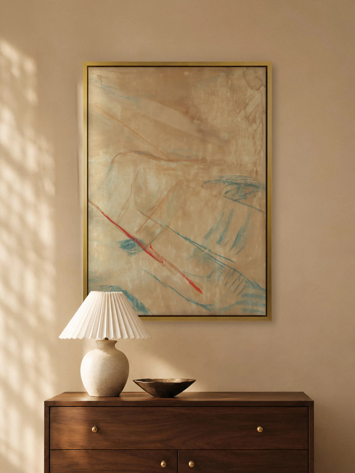Abstraktes Leinwandbild in Beige, Blau und Rot im Goldrahmen über einer Kommode aus dunklem Holz mit Keramiklampe und Metallschale. Das Japandi Wandbild greift das warme Licht des Raums auf.