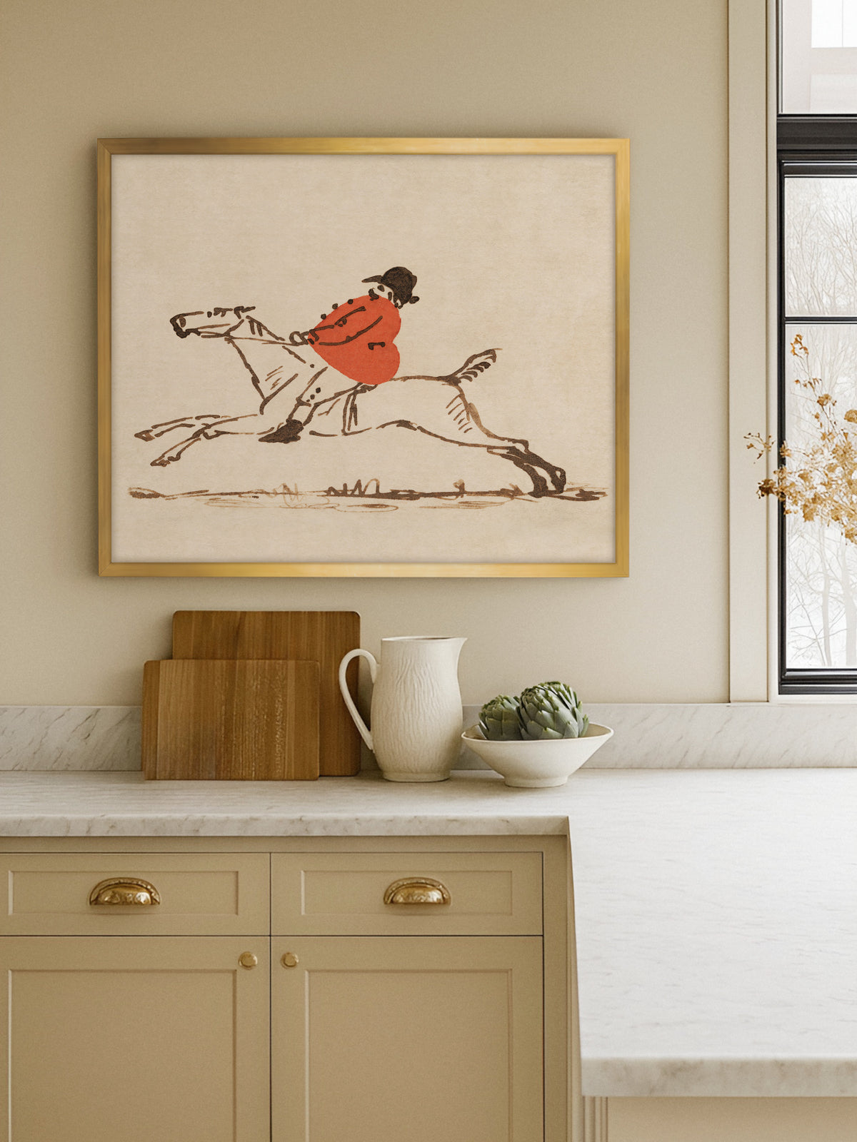 Vintage Pferde Wandbild im goldenen Rahmen in einer beigen Küche über einer Marmorarbeitsplatte mit Holzbrettern, weißem Krug und Artischocken in einer Schale. Die Reiterskizze setzt einen warmen roten Akzent neben dem Fenster.
