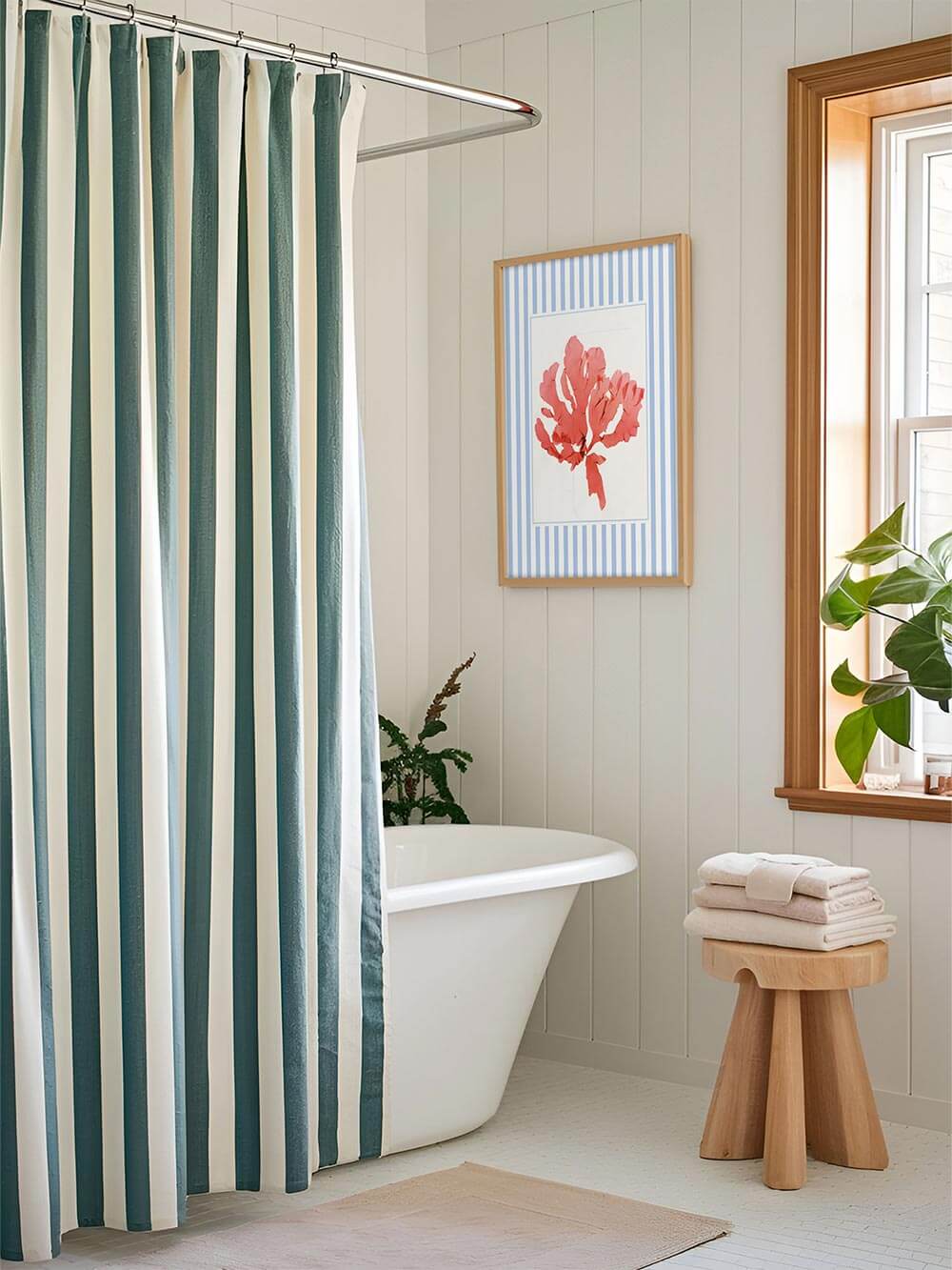 Der Algen-Kunstdruck mit blauer Umrahmung hängt neben der Badewanne. Die botanische Illustration harmoniert mit den gestreiften Vorhängen und dem hellen Interieur.