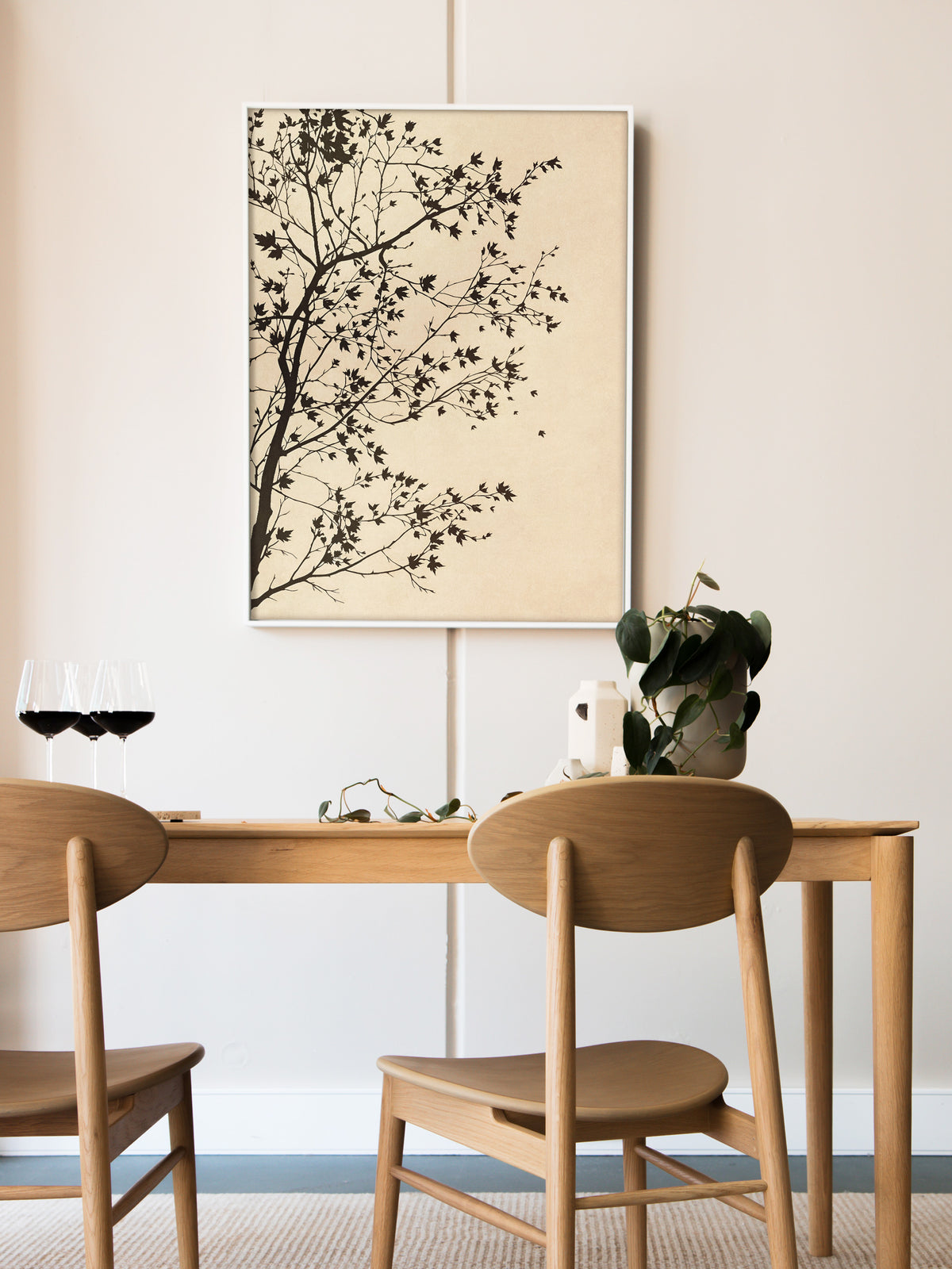 Kunstdruck mit Baumsilhouette im weißen Rahmen über einem schlichten Esstisch aus hellem Holz mit zwei Holzstühlen, Rotweingläsern und einer Grünpflanze. Die skandinavisch helle Einrichtung lässt die dunklen Zweige klar hervortreten.