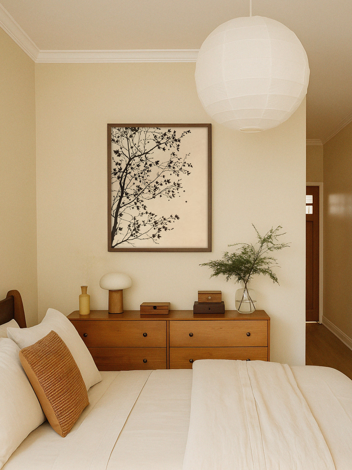 Wandbild mit dunklen Ahornzweigen im Rahmen aus dunklem Holz über einer Holzkommode mit Pilzlampe und Glasvase, davor ein Bett mit heller Leinenwäsche. Eine runde Papierleuchte unterstreicht den Japandi Stil des Schlafzimmers.