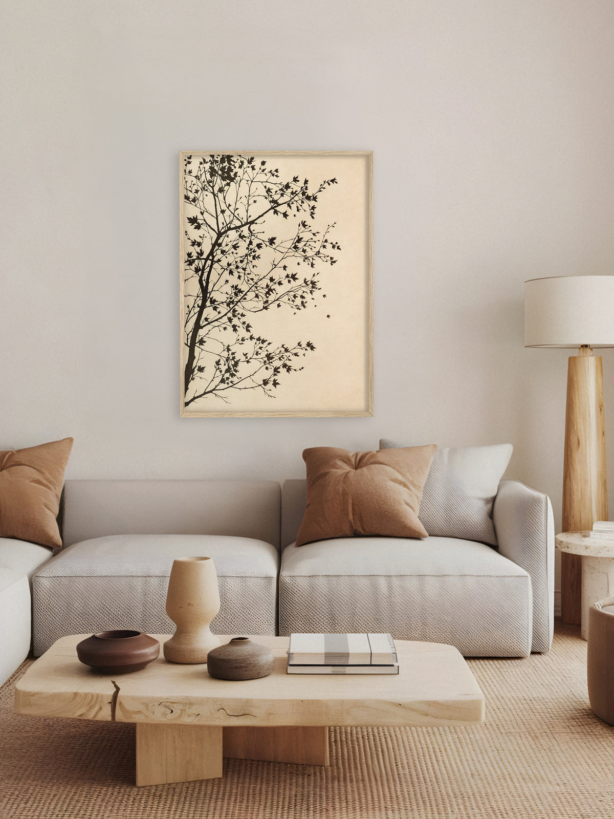 Poster mit Baumsilhouette im Rahmen aus hellem Holz über einem hellgrauen Sofa mit braunen Kissen, davor ein massiver Holztisch mit Keramikvasen. Die Stehleuchte aus Holz und die Sandtöne ergeben ein ruhiges Japandi Wohnzimmer.