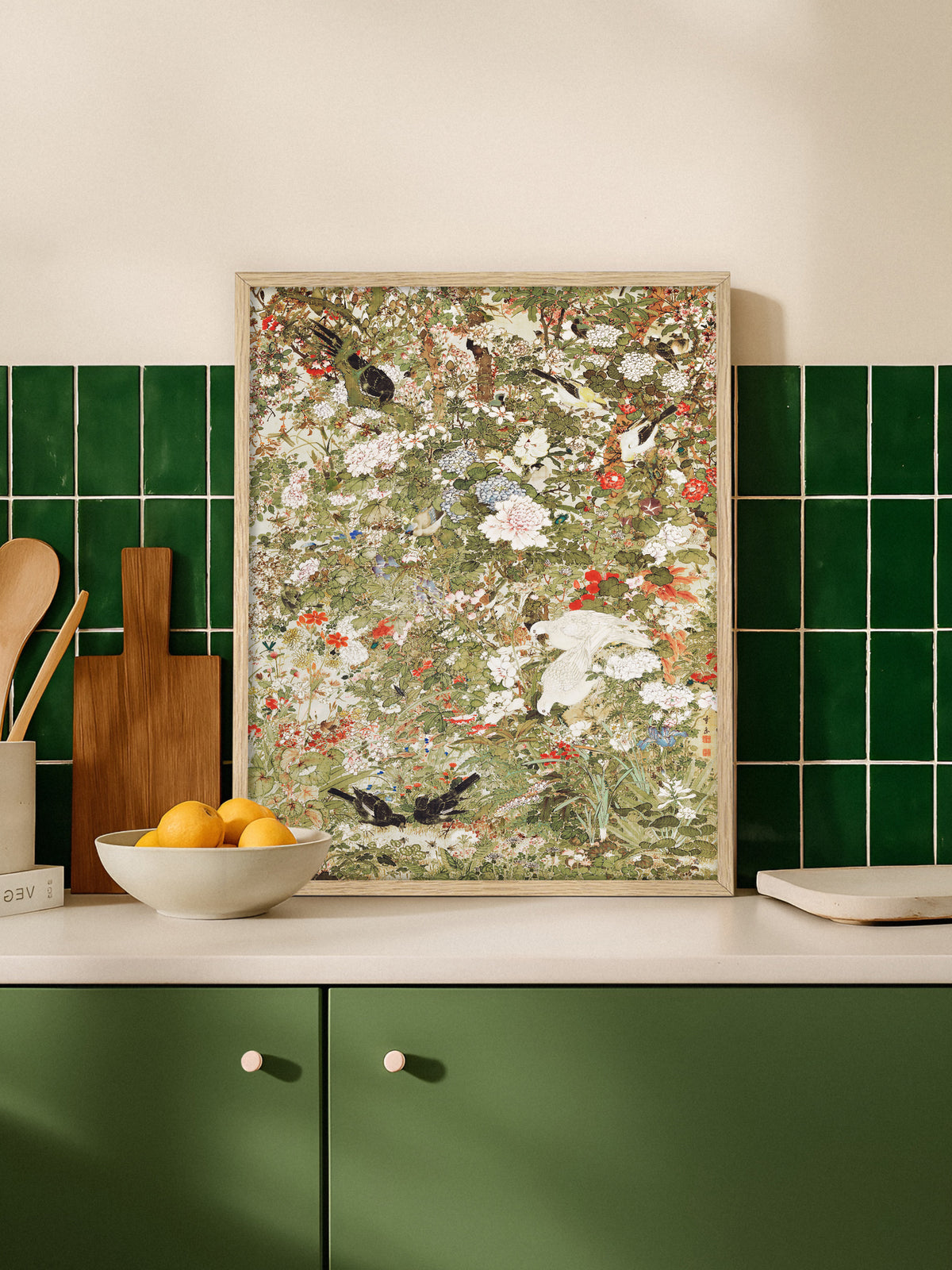Poster mit Vögeln und Blumen im Rahmen aus hellem Holz, angelehnt auf einer Küchenarbeitsplatte vor dunkelgrünen Fliesen, daneben Schneidebrett und eine Schale mit Orangen. Das Grün des Motivs greift die Fliesen und Schrankfronten auf.