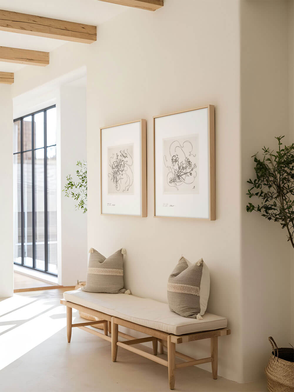 Zwei schlichte, gerahmte Kunstposter mit floralen Schwarz-Weiß-Zeichnungen hängen nebeneinander an einer hellen, minimalistischen Flurwand über einer schlichten Holzbank mit zwei beigen Kissen.