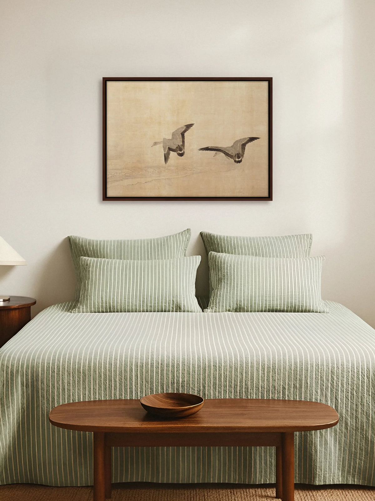 Japanisches Vogel Leinwandbild im braunen Rahmen über einem Bett mit salbeigrün gestreifter Bettwäsche. Am Fußende steht eine ovale Holzbank mit einer Schale, die Wand dahinter ist hell und schlicht.
