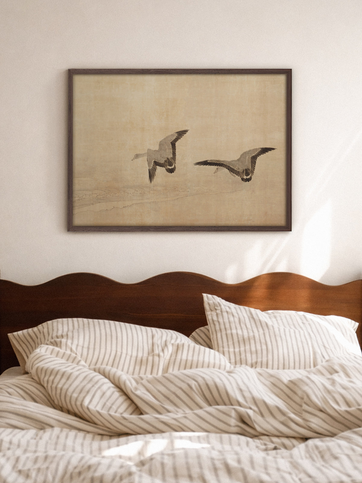 Kunstdruck mit zwei fliegenden Gänsen im Rahmen aus dunklem Holz über einem Bett mit gewelltem Kopfteil aus Holz und gestreifter Bettwäsche. Das sandfarbene Wabi-Sabi-Wandbild bringt Ruhe ins helle Schlafzimmer.