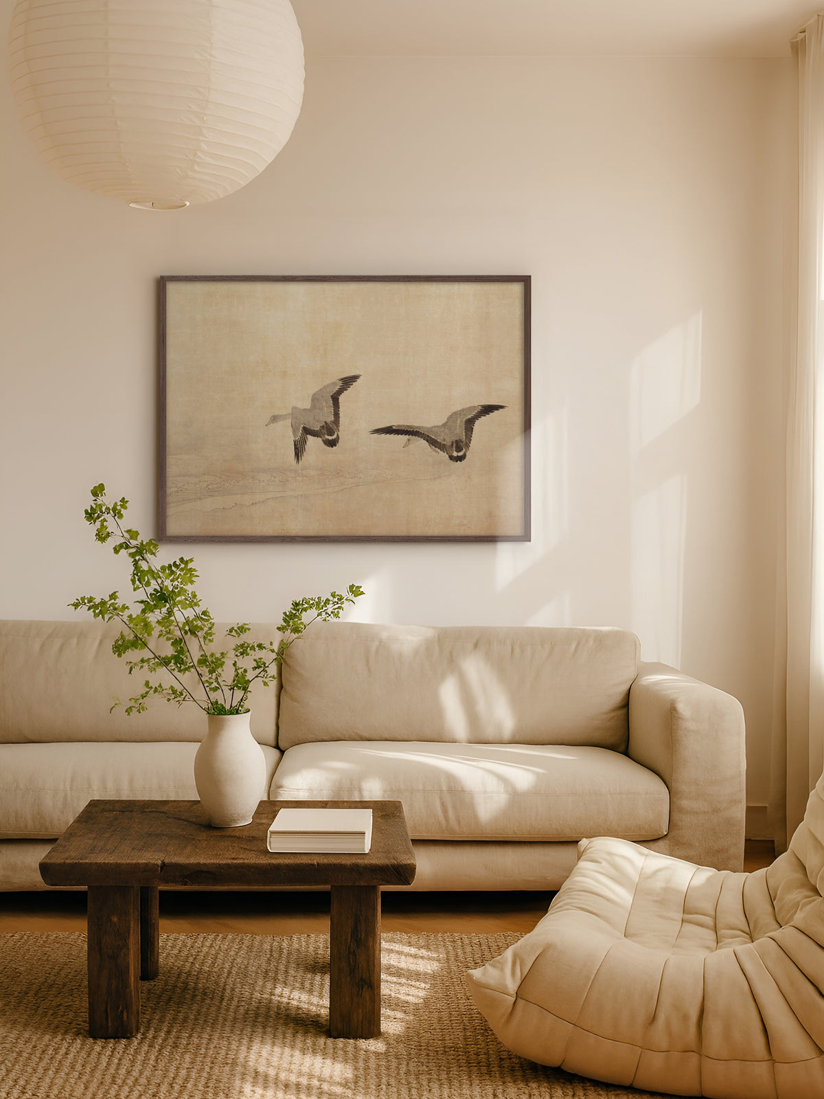 Poster mit zwei fliegenden Gänsen im Rahmen aus dunklem Holz über einem hellen Leinensofa, davor ein rustikaler Holztisch, darüber eine Papierleuchte im warmen Sonnenlicht. Das Wandbild im Japandi-Stil passt zu den Naturtönen des Wohnzimmers.