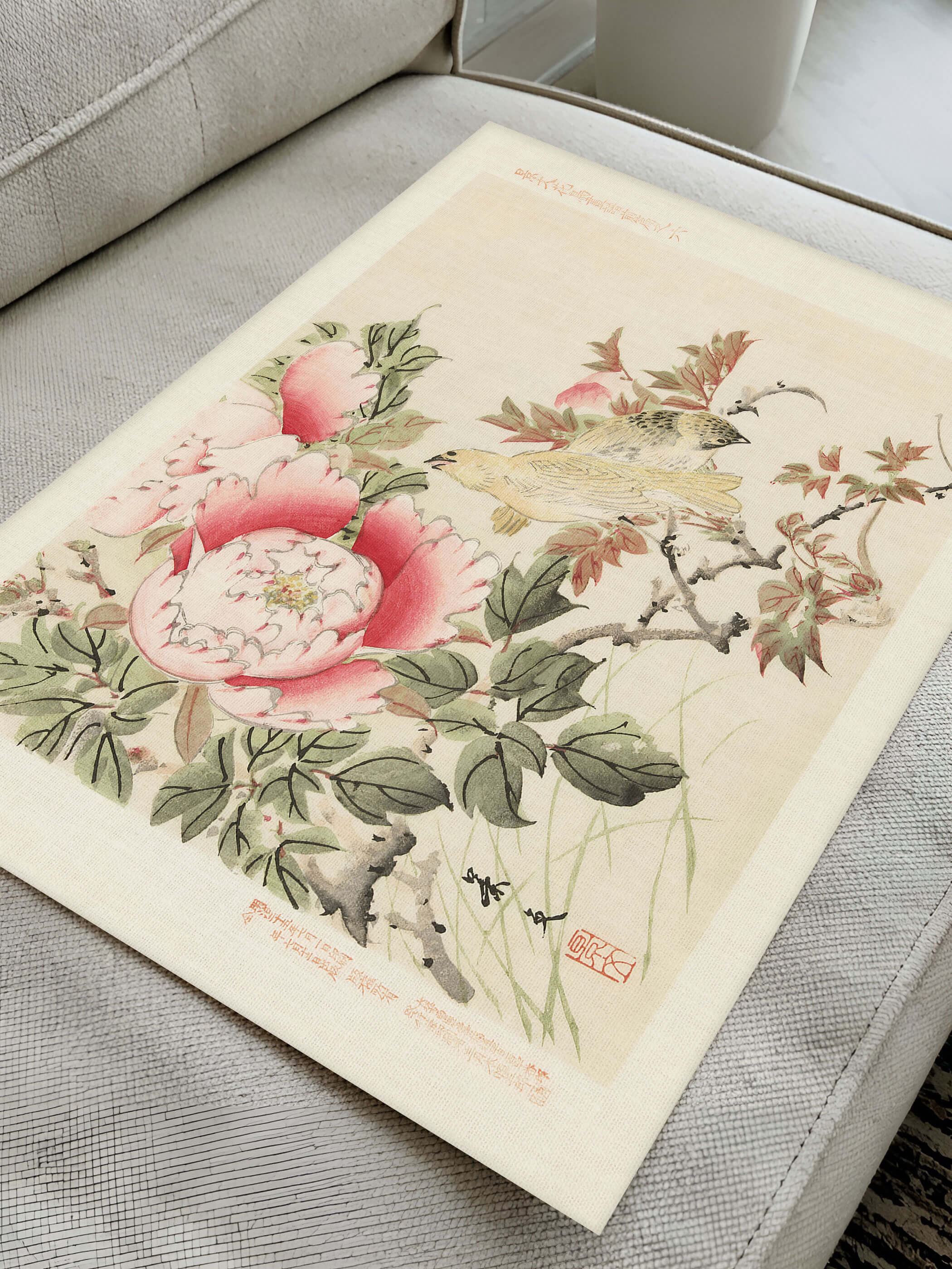 Flatlay des Chinoiserie Kunstdrucks auf einem Sofa liegend zeigt die Details der Vögel und Pfingstrosenblüten in warmen Rosa- und Grüntönen. Der rote Siegelstempel betont den vintage Charakter des Wandbilds.