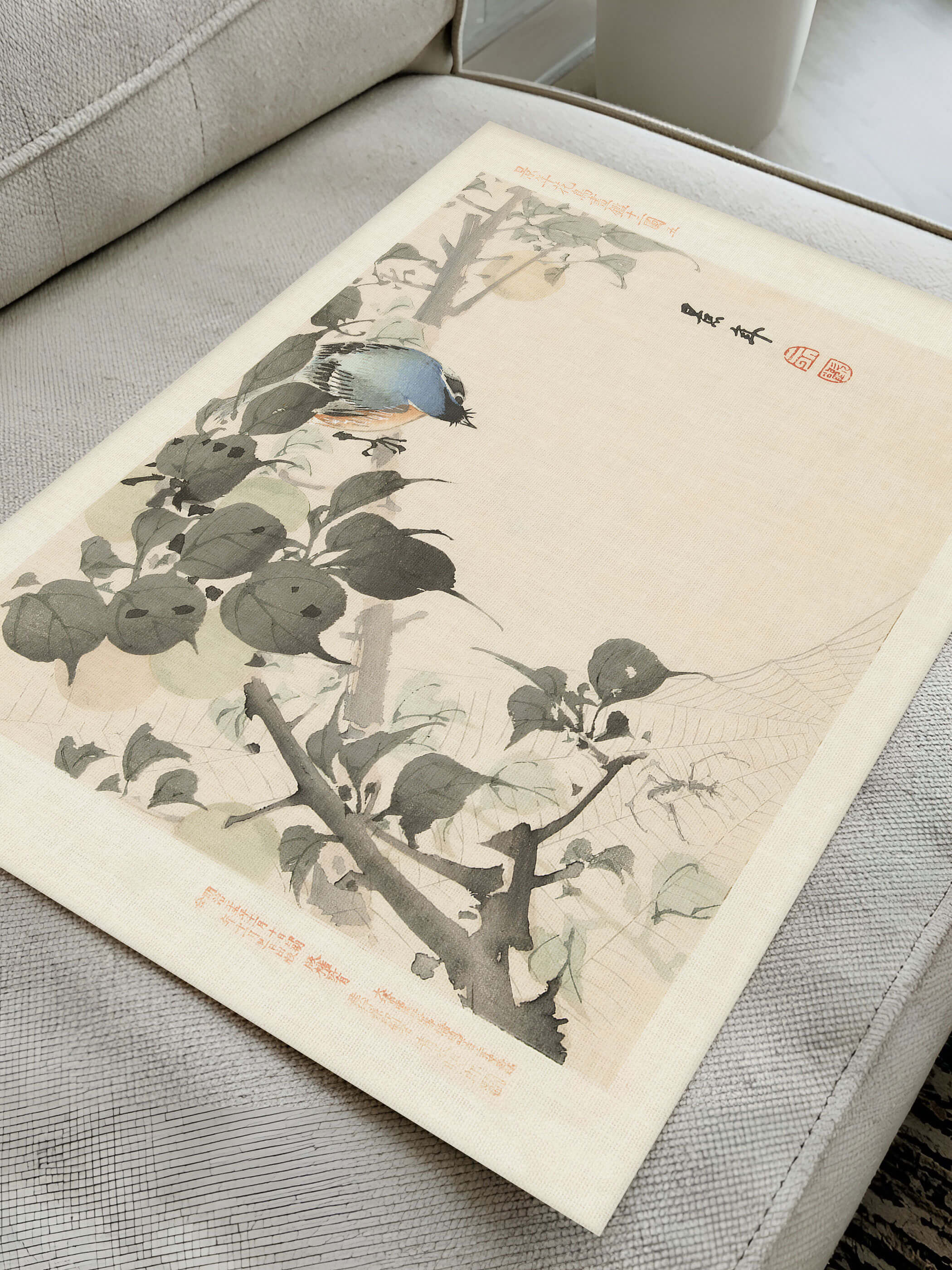 Flatlay des Chinoiserie Kunstdrucks auf einem Sofa liegend zeigt einen blau-orangefarbenen Vogel zwischen grau getönten Blättern im Detail. Der rote Siegelstempel betont den vintage Charakter des Wandbilds.