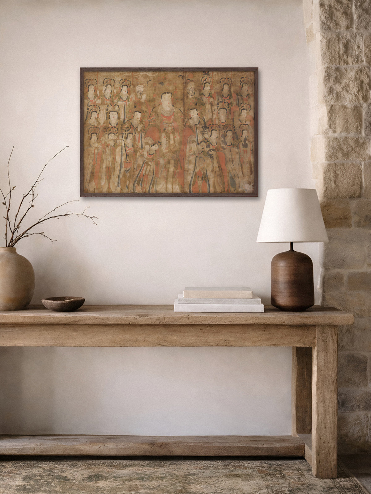 Vintage Kunstdruck mit asiatischer Figurenszene im dunklen Holzrahmen über einer rustikalen Holzkonsole mit Tischlampe, Bücherstapel, Holzschale und Vase mit Zweigen neben einer Natursteinwand. Das Poster passt zu einer ruhigen Wabi-Sabi Einrichtung.