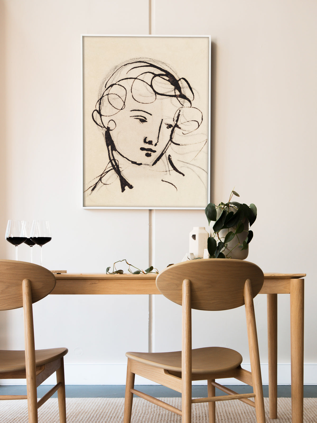 Wandbild mit der Tuscheskizze eines Frauenkopfs im weißen Rahmen über einem Esstisch aus Eiche mit Rotweingläsern und Grünpflanze. Die reduzierten Linien passen zu der ruhigen, minimalistischen Einrichtung.