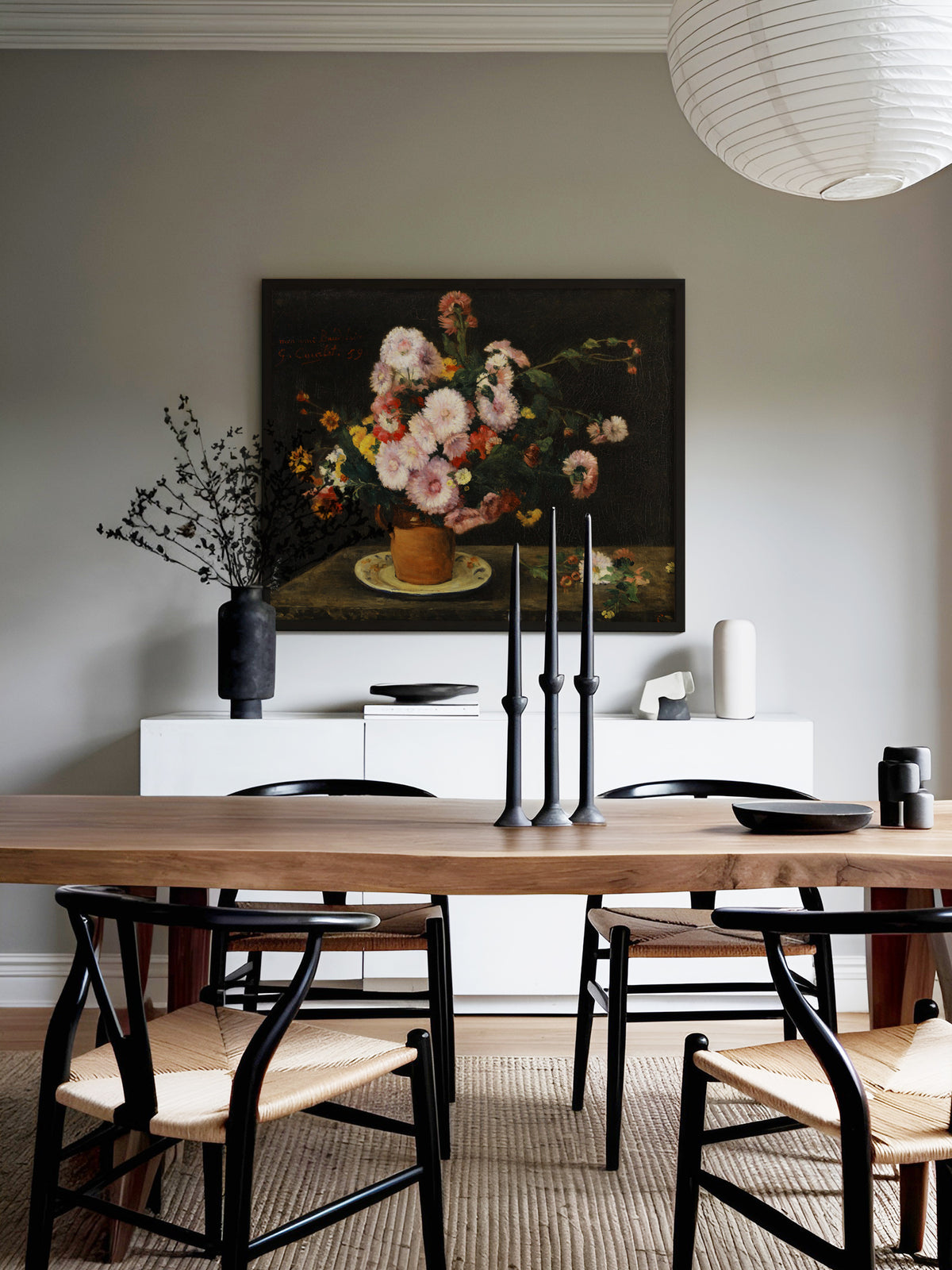 Dunkles Blumenstillleben im schwarzen Rahmen über einem weißen Sideboard im Esszimmer, davor ein Holztisch mit schwarzen Kerzenleuchtern und Stühlen mit Geflechtsitz unter einer Papierlampe. Der Kunstdruck setzt einen farbigen Akzent vor der grauen Wand.