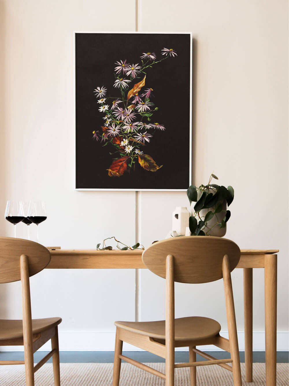 Ein filigraner Kunstdruck mit pastellfarbenen Blumen auf schwarzem Hintergrund hängt in einem minimalistischen Esszimmer. Die zarten, weiß-rosa Blüten heben sich lebendig vom dunklen Hintergrund ab. Der helle Holztisch und die beiden Weingläser im Vordergrund verleihen dem Bild eine warme, einladende Atmosphäre.