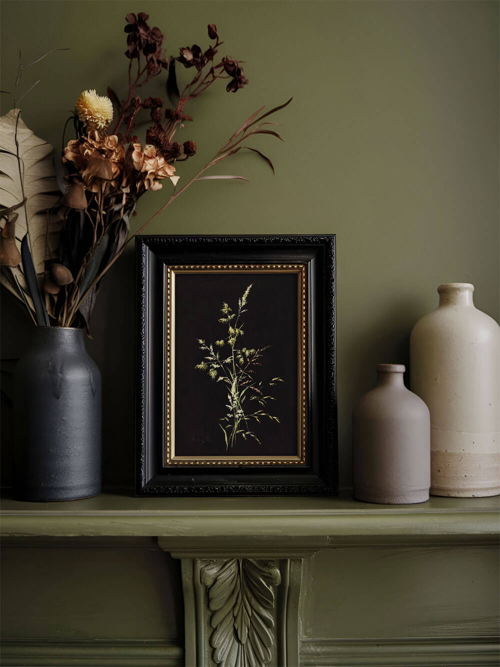 Ein botanischer Kunstdruck, eingerahmt in einem schwarzen Rahmen mit goldener Innenkante, steht auf einem grünen Kaminsims. Das Bild zeigt einen filigranen Grashalm auf dunklem Hintergrund. Umgeben wird es von getrockneten Blumen in warmen Brauntönen und mattierten Keramikvasen in gedeckten Naturfarben. Die Szene strahlt eine ruhige, nostalgische Atmosphäre aus, betont durch den gedämpften Grünton der Wand.