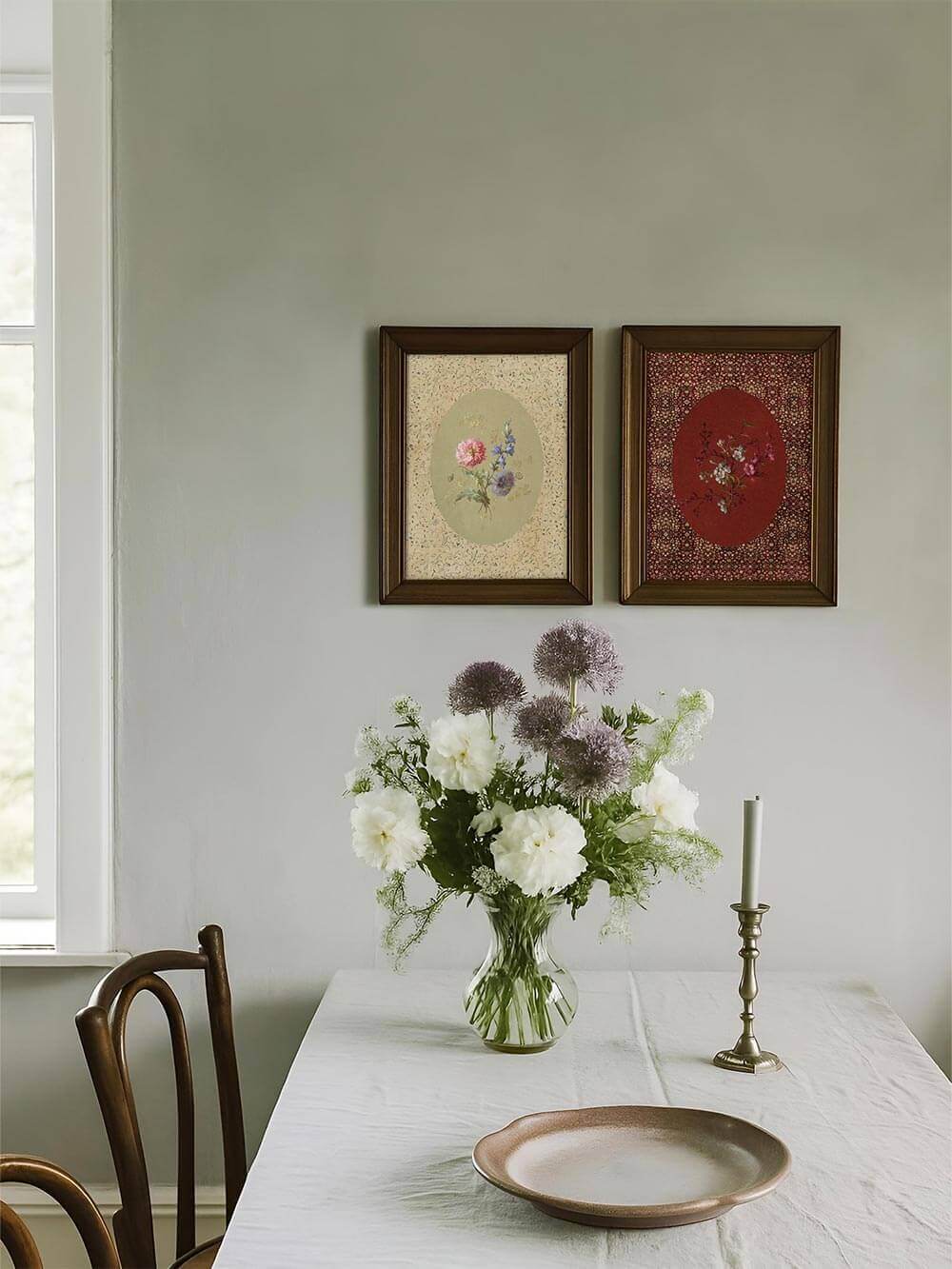 Der florale Kunstdruck mit ovalem Design hängt neben einem roten Pendant über einem schlichten Esstisch mit Wildblumenstrauß und perfekt für französisch inspirierte Essbereiche.