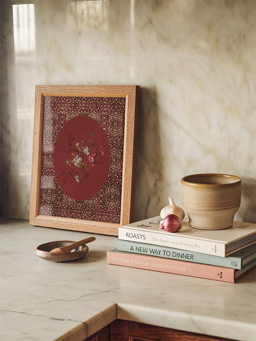 Ein gerahmter Blüten-Kunstdruck lehnt an einer Küchenwand aus Marmor. Der rote Hintergrund und das zarte florale Motiv setzen einen dekorativen Akzent zwischen Kochbüchern und Keramik.