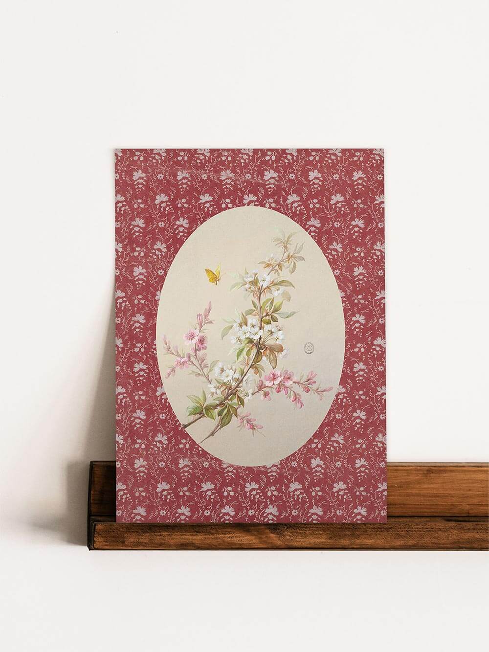 Der botanische Kunstdruck lehnt auf einer schlichten Holzleiste an der Wand. Feine Blüten, ein Schmetterling und der gemusterte rote Rahmen verleihen ihm einen nostalgischen Charme.
