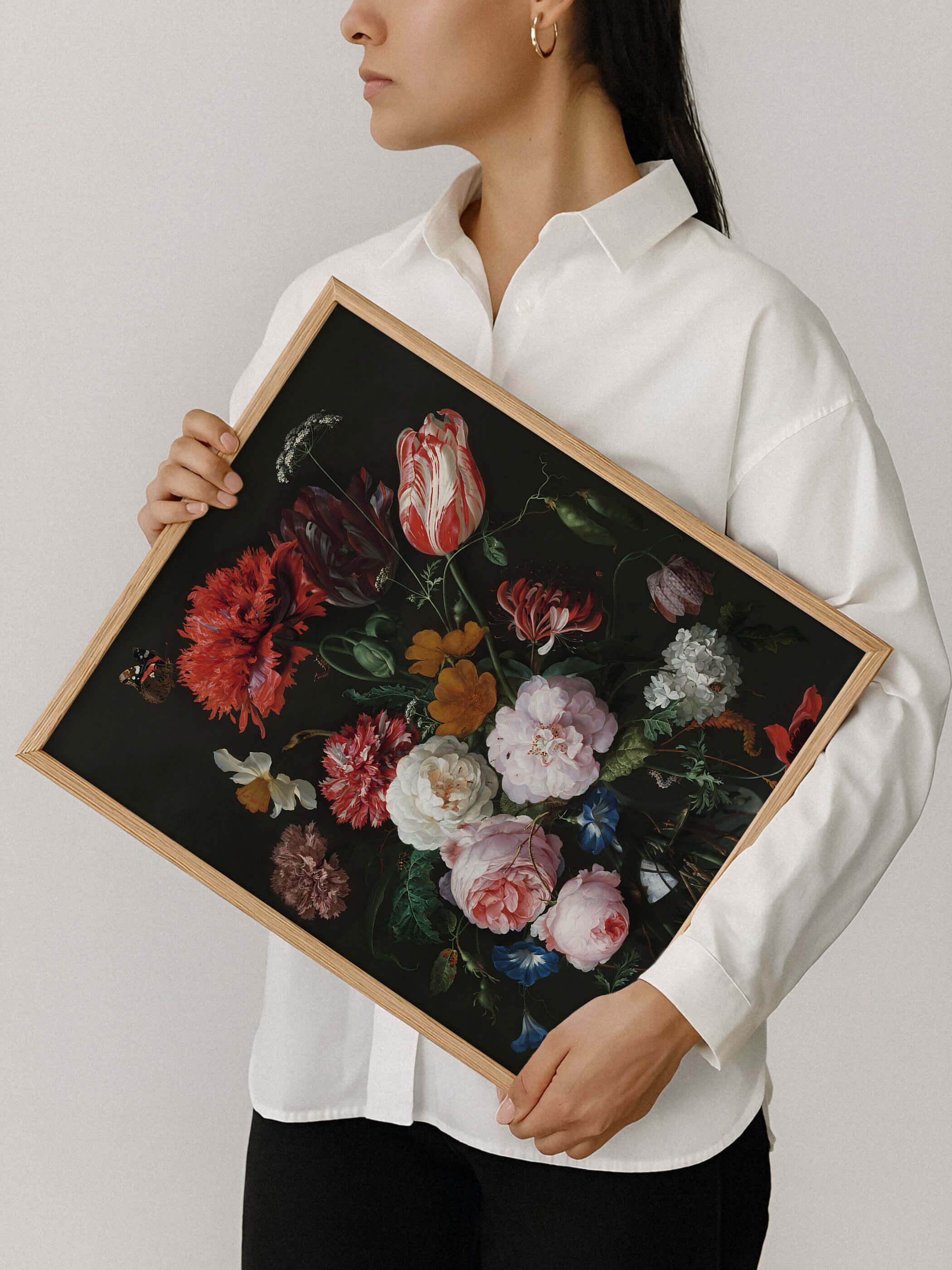 Eine Frau in weißem Hemd hält einen Holzrahmen mit dem barocken Blumenstillleben aus Mohn und Rosen vor neutralem Hintergrund. Der Kunstdruck zeigt die kräftigen Farben und die feine Rahmung im Detail.