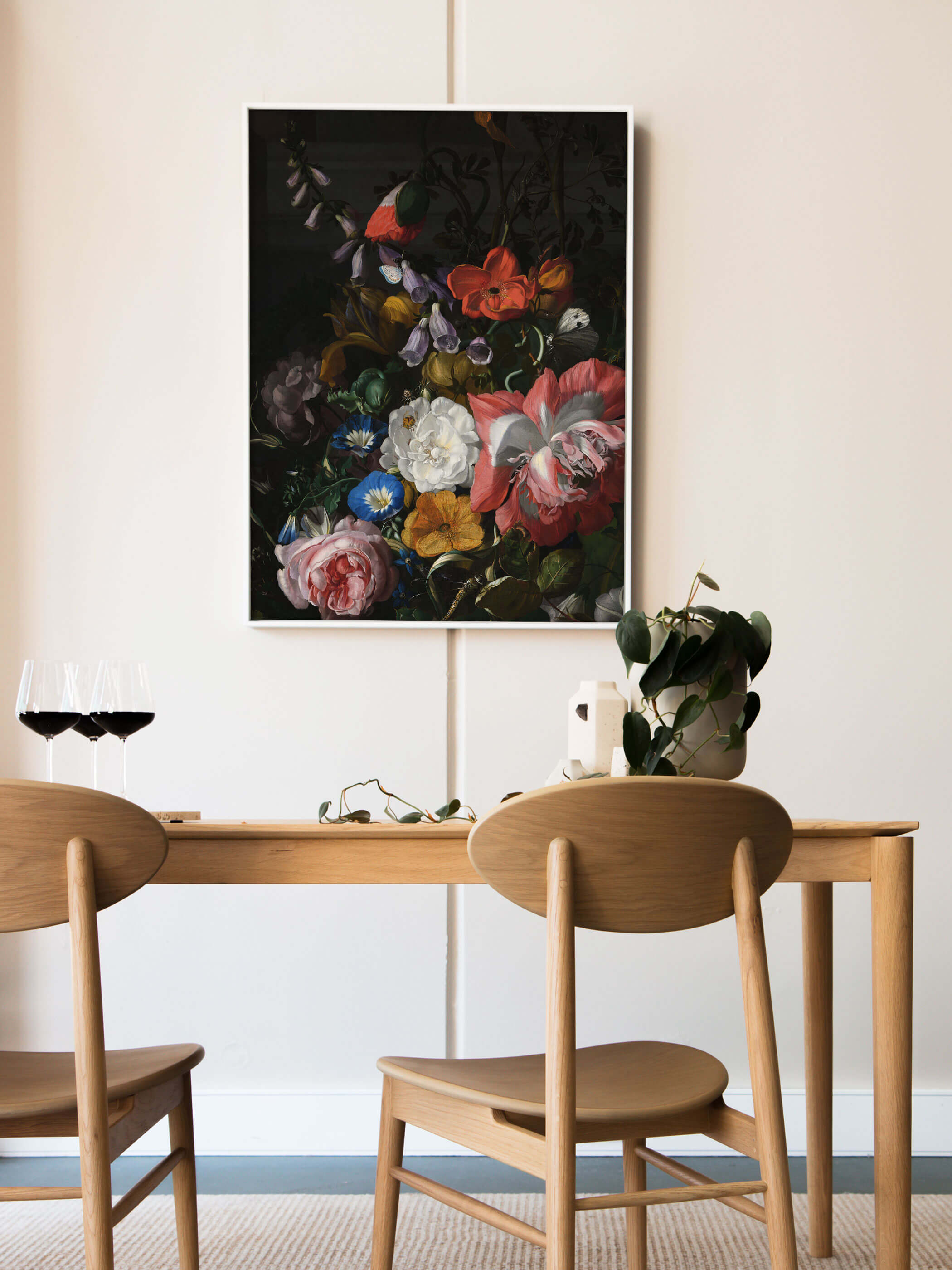 Weiß gerahmter Kunstdruck mit barockem Blumenstillleben lehnt an der Wand über einem Holztisch mit Rotweingläsern und einer grünen Pflanze. Das dunkle, farbintensive Motiv setzt einen eleganten Akzent im hellen Esszimmer.