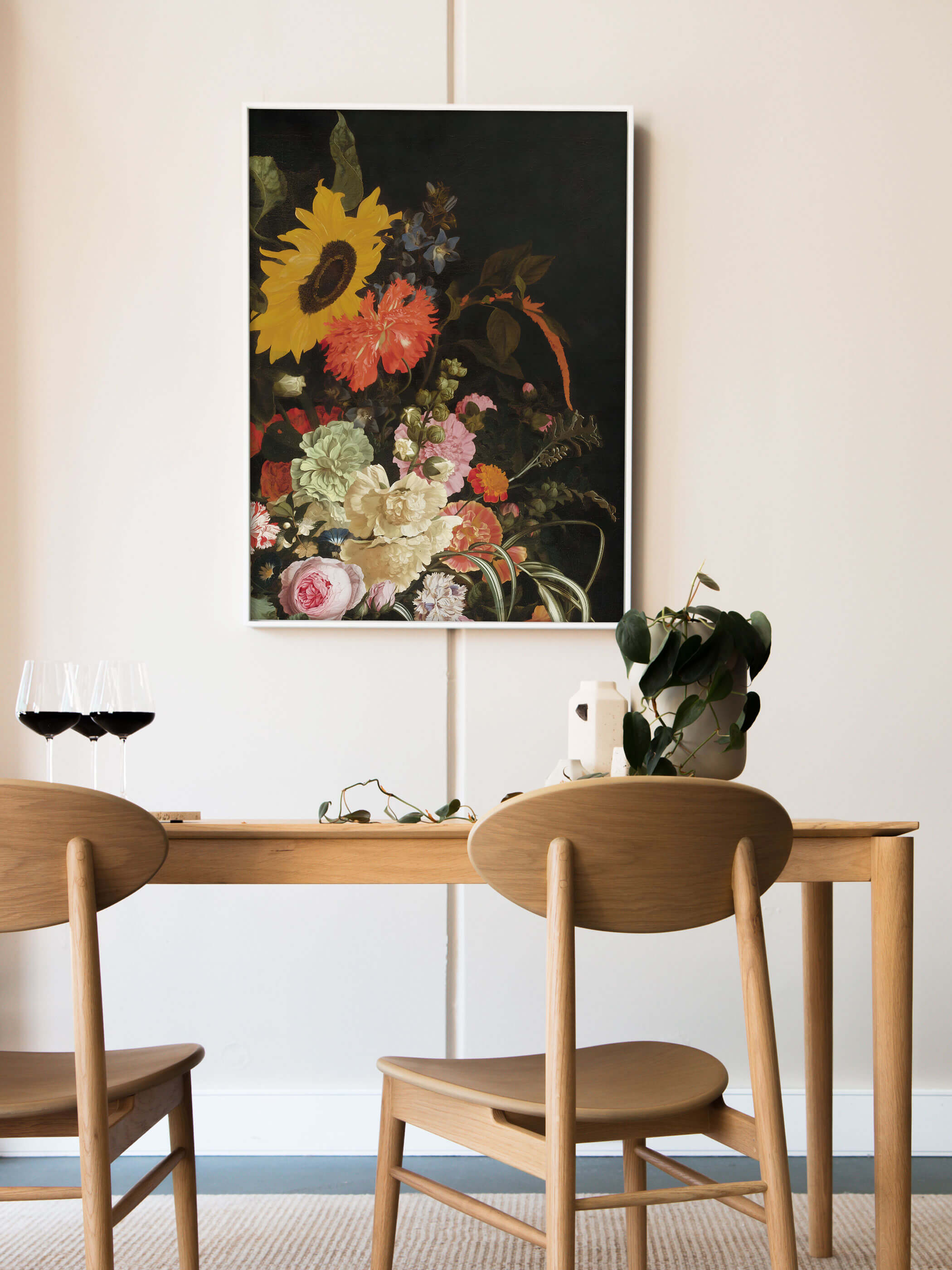 Weiß gerahmter Kunstdruck mit barockem Blumenstillleben lehnt an der Wand über einem Holztisch mit Rotweingläsern und einer grünen Pflanze. Das dunkle, farbenfrohe Motiv setzt einen eleganten Akzent im hellen Esszimmer.
