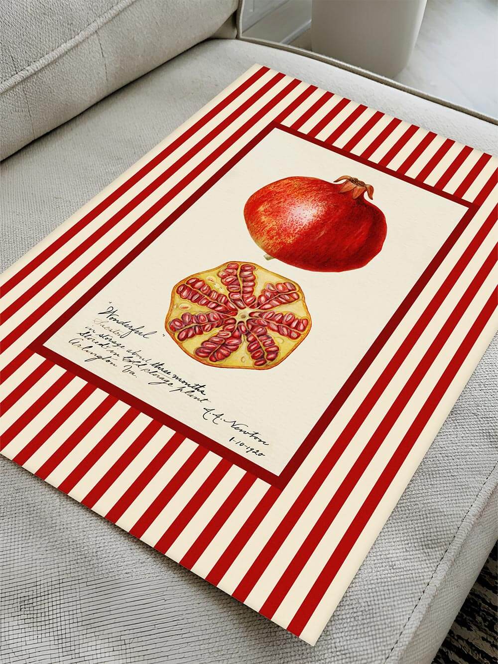 Vintage Granatapfel-Kunstdruck mit auffälligem rot-weißem Streifenrahmen, flach auf Sofa drapiert – zeigt botanisches Fruchtmotiv mit handschriftlichem Text, ideal für stilvolle Wandgestaltung.