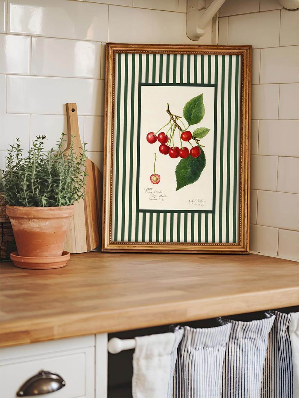 Vintage-Kirsch-Kunstdruck im goldenen Rahmen auf Küchenarbeitsplatte mit weiß gefliester Wand, Holzdetails und Kräutertopf – französischer Landhausstil.