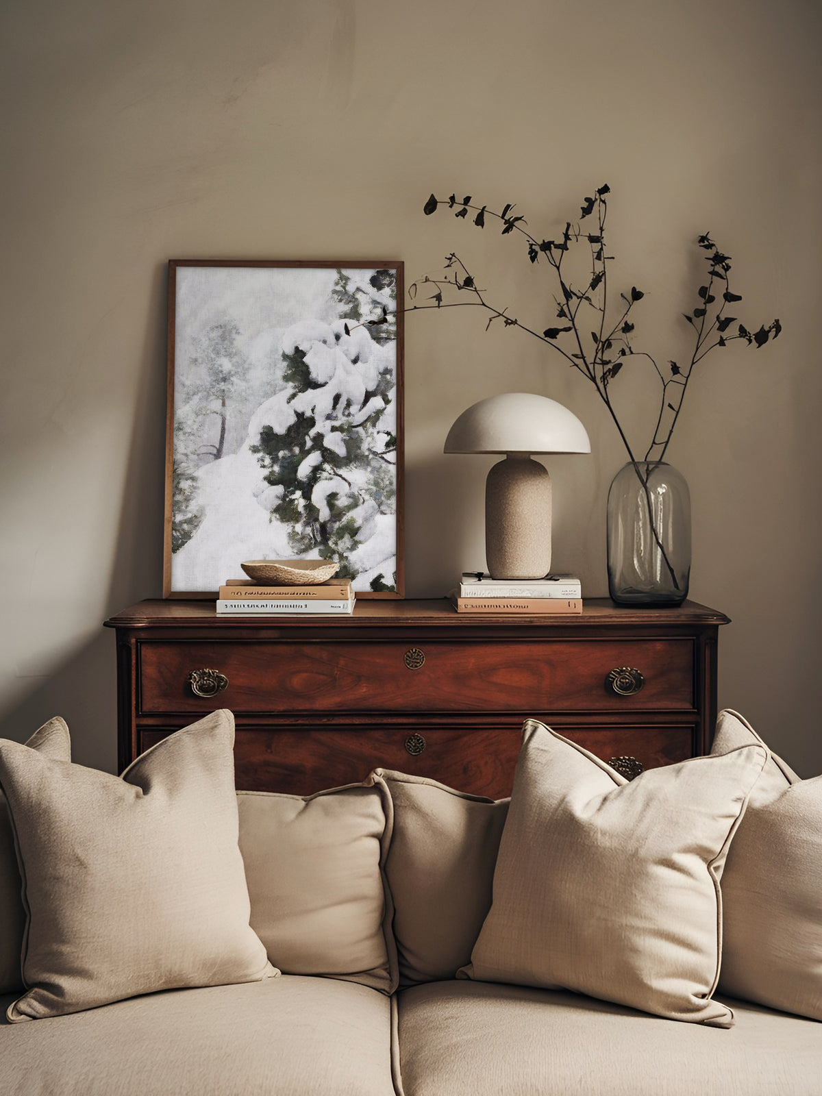 Wabi Sabi Wandbild einer Winterlandschaft im dunklen Holzrahmen, angelehnt auf einer antiken Kommode mit Pilzlampe und Glasvase vor einem beigen Sofa im Wohnzimmer.