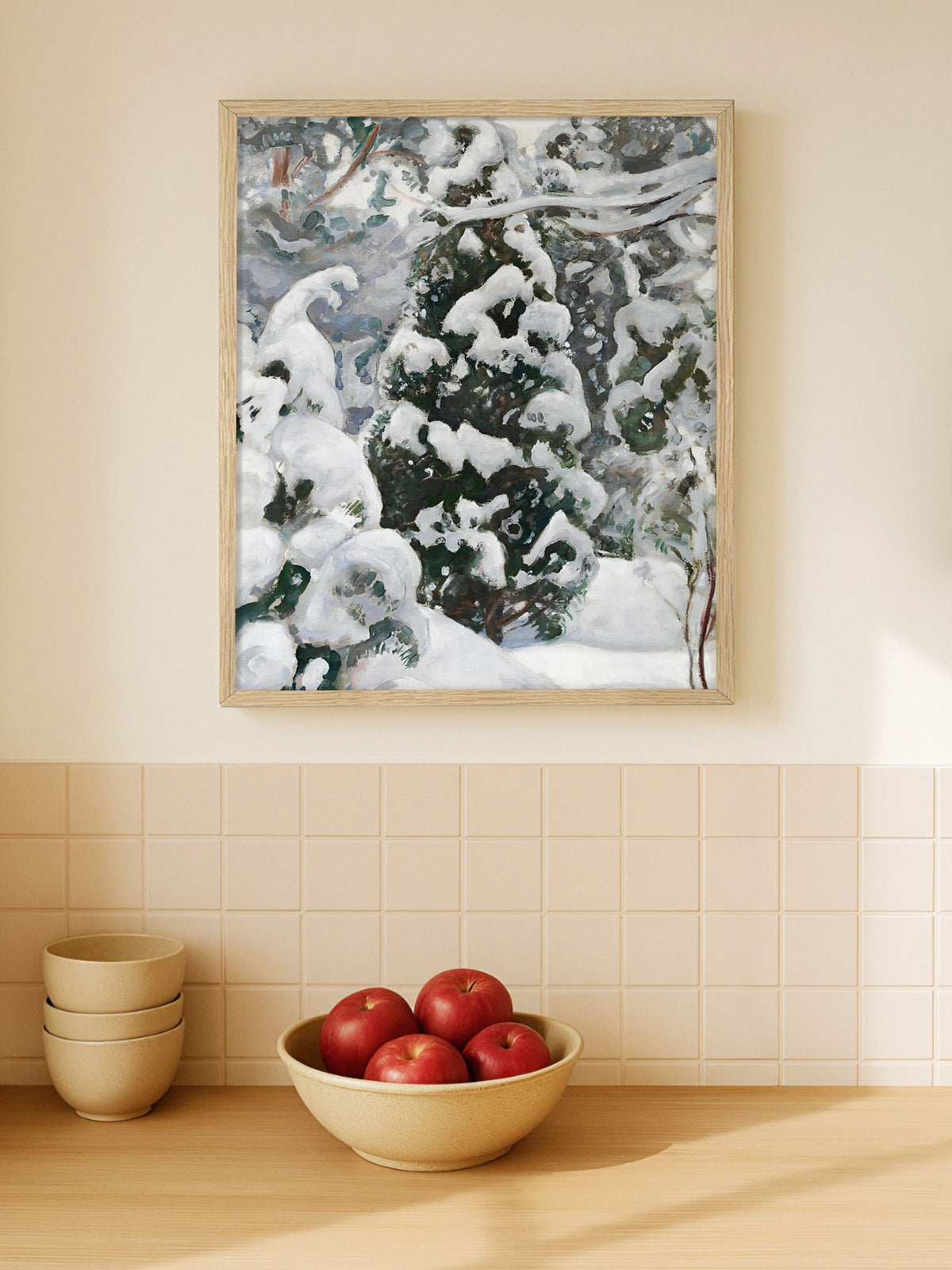 Winterliches Poster mit verschneitem Wacholder im hellen Holzrahmen in einer hellen Küche über gefliester Rückwand, davor Schalen und rote Äpfel auf der Arbeitsfläche.