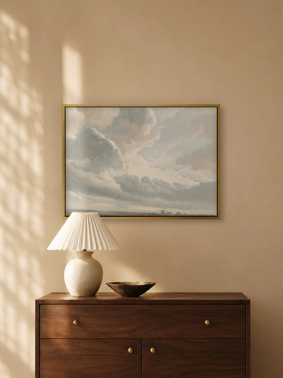 Pastellfarbenes Wolkenbild auf Leinwand im goldenen Rahmen über einer dunklen Holzkommode mit Messingknöpfen. Auf der Kommode stehen eine Keramiklampe mit weißem Plisseeschirm und eine flache Bronzeschale.