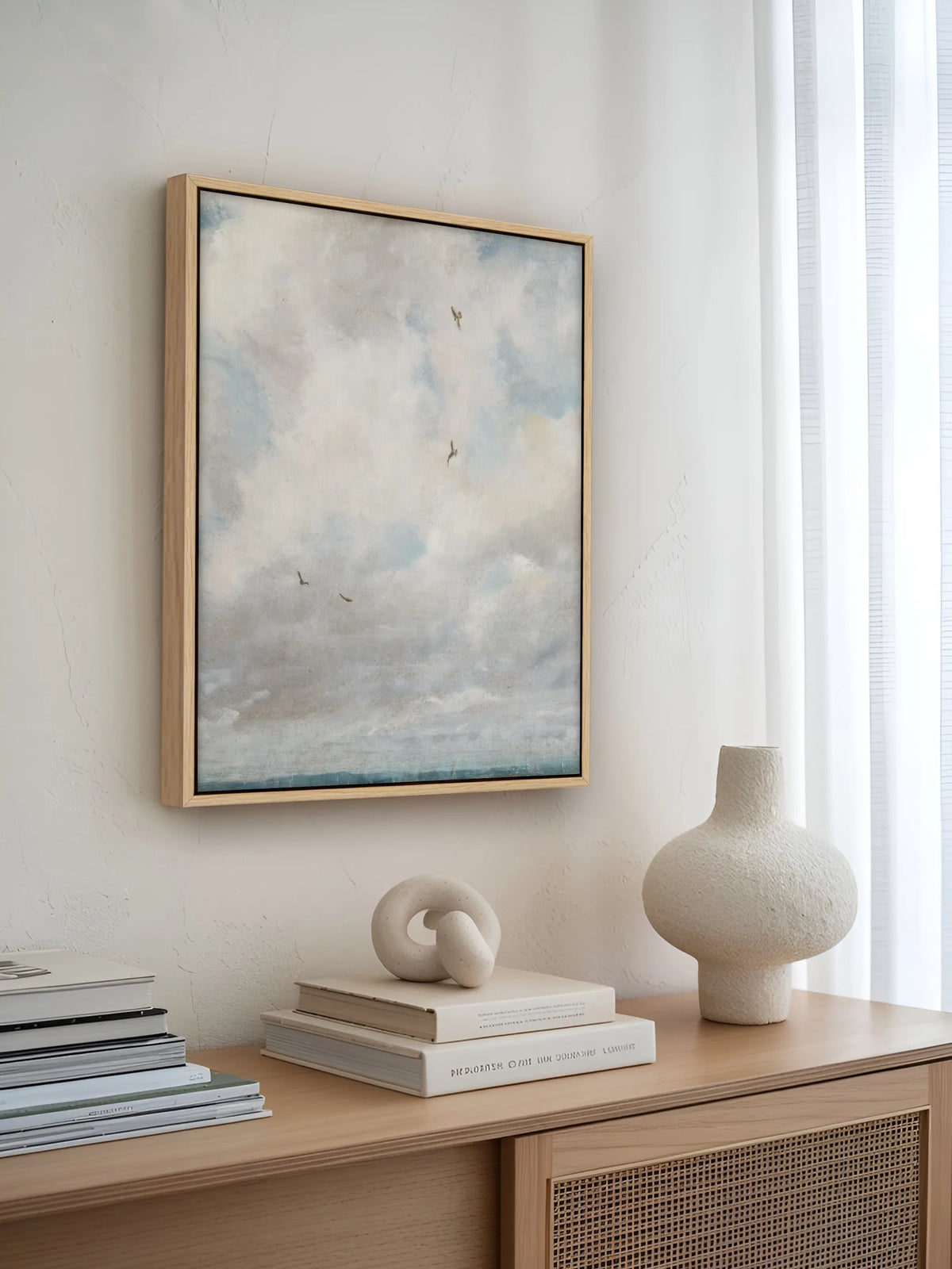 Himmel Leinwandbild im Naturholzrahmen an einer weißen Putzwand über einem hellen Sideboard mit Rattanfront. Darauf stehen eine cremefarbene Keramikvase, eine Knotenskulptur auf Büchern und ein Stapel Magazine.