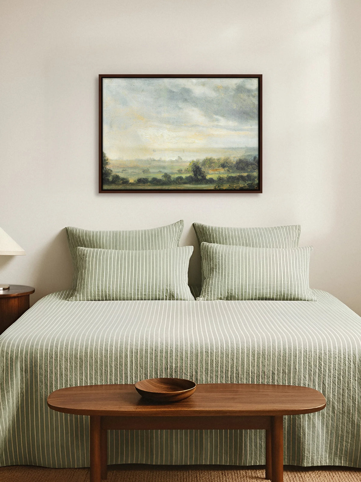 Vintage Landschaft als Leinwandbild im braunen Rahmen über einem Bett mit salbeigrün gestreifter Leinenbettwäsche. Am Fußende steht eine ovale Holzbank mit einer Holzschale, die Wand ist cremeweiß.