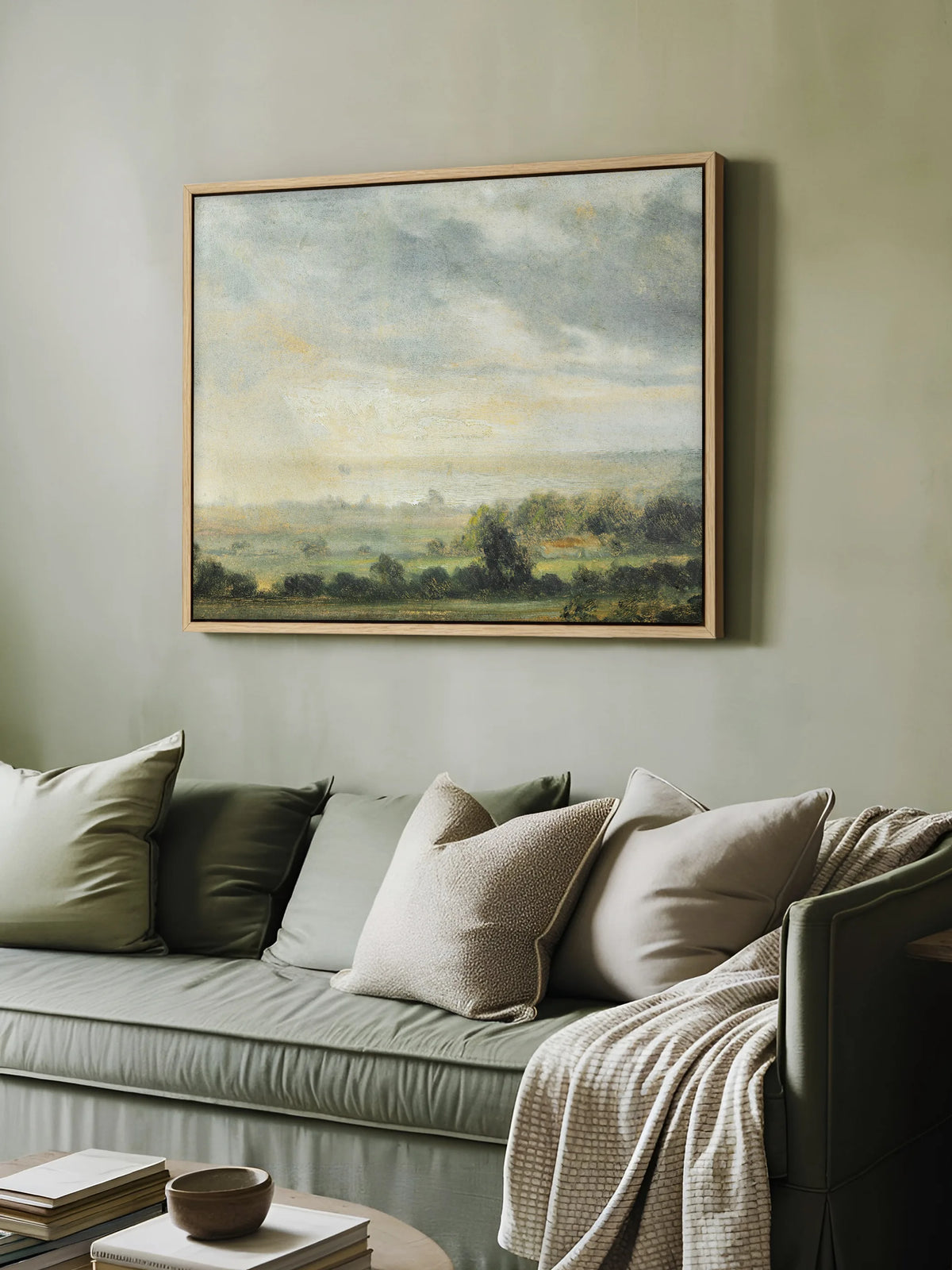 Das Cottagecore Leinwandbild mit weiter Landschaft hängt im Naturholzrahmen an einer salbeigrünen Wand über einem grünen Sofa. Auf dem Sofa liegen Kissen in Grün und Beige und eine cremefarbene Strickdecke.