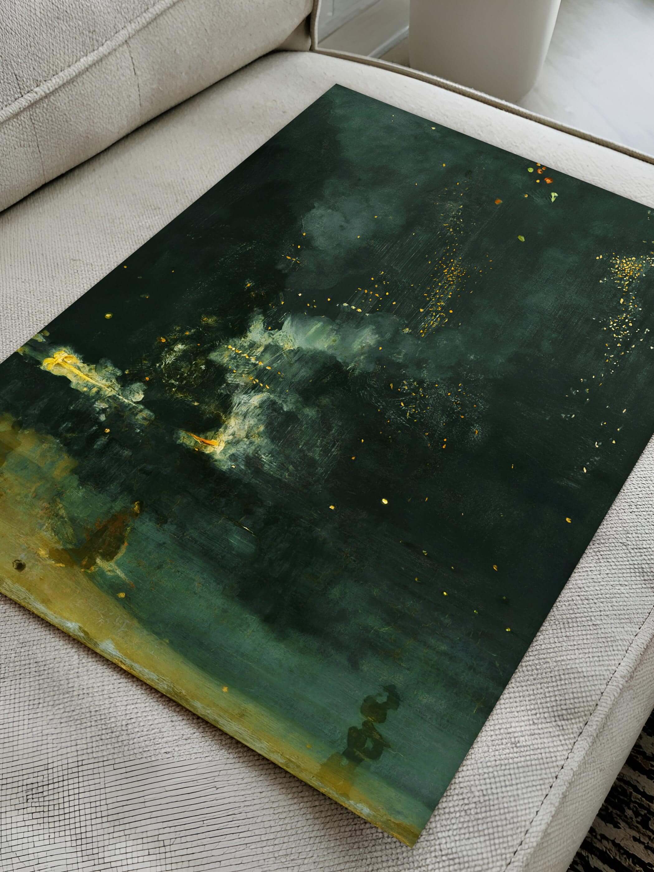 Detailansicht des Kunstdrucks "Nocturne in Black and Gold" von James Abbott McNeill Whistler, flach auf einem Sofa liegend. Der Druck zeigt eine düstere Nachtszene mit goldenen Funken und Licht.