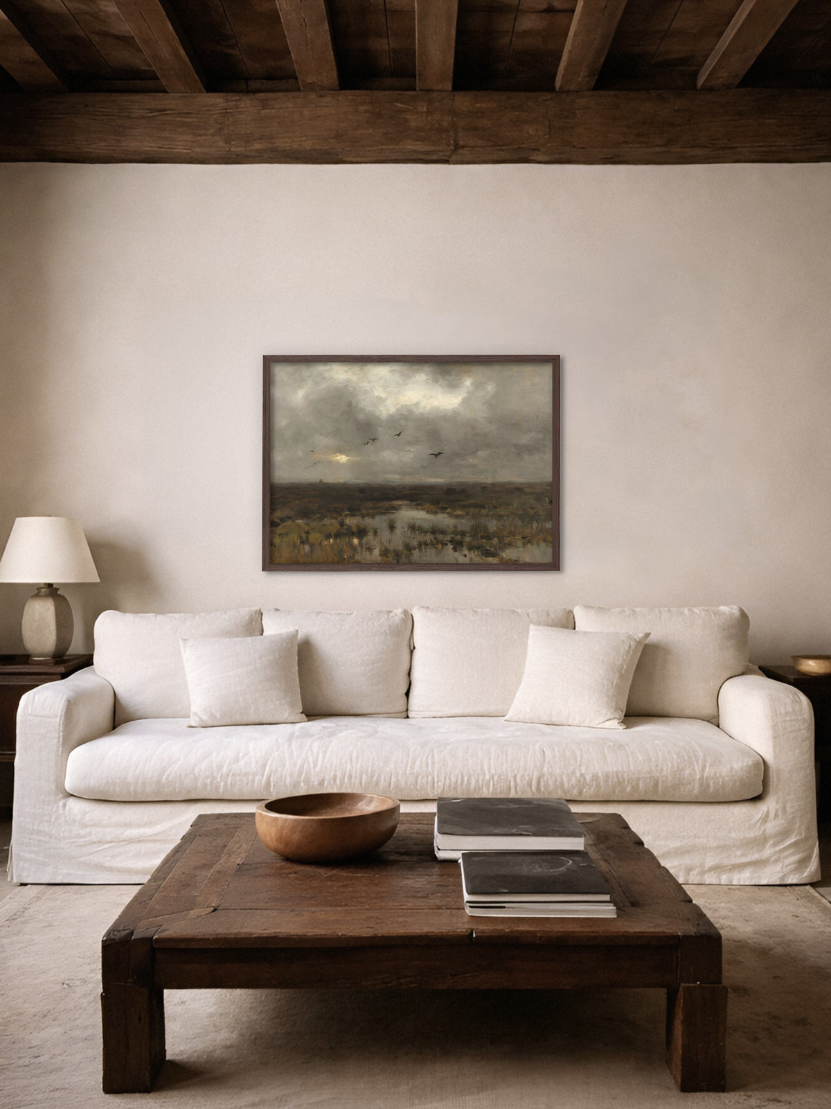 Wabi Sabi Wandbild einer Moorlandschaft im dunklen Holzrahmen über einem cremefarbenen Leinensofa, davor ein rustikaler Couchtisch mit Holzschale, darüber dunkle Deckenbalken.
