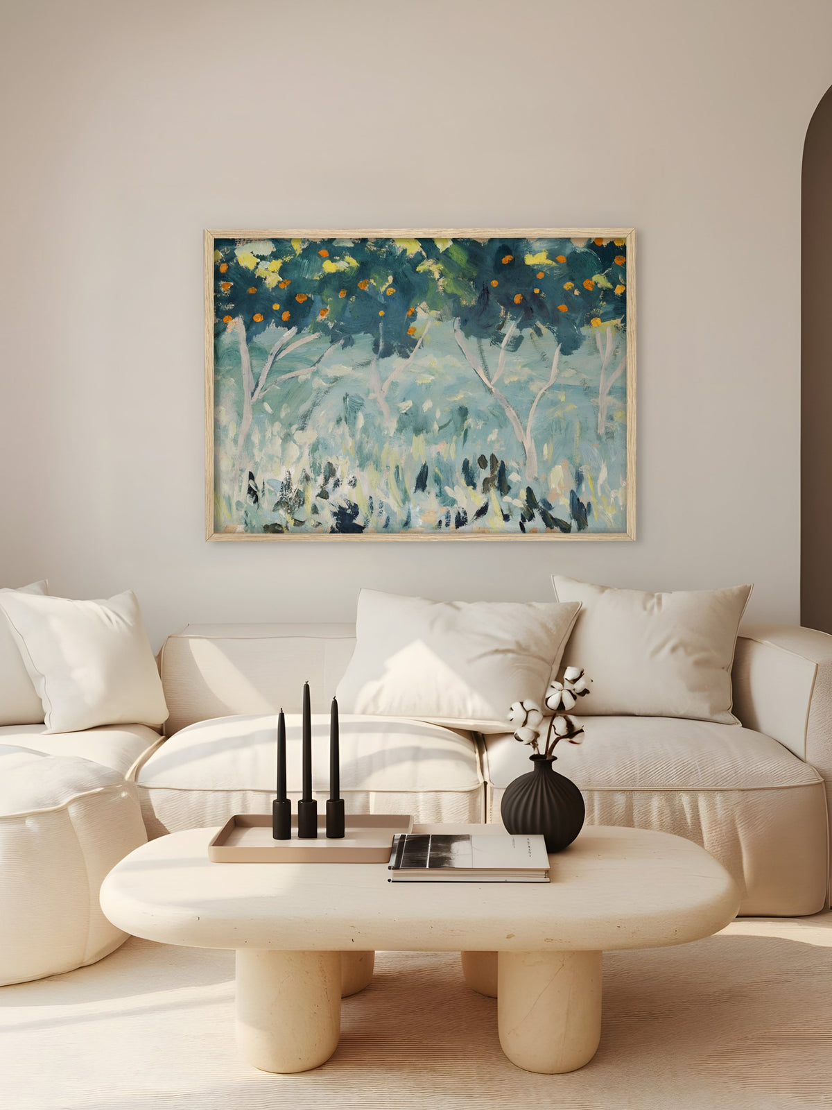 Kunstdruck mit Orangenbäumen im hellen Holzrahmen über einem cremefarbenen Sofa mit hellen Kissen, davor ein ovaler Couchtisch aus Stein mit schwarzen Kerzen und einer schwarzen Vase mit Baumwollzweigen. Das mediterrane Wandbild setzt einen farbigen Akzent im beigen Wohnzimmer.