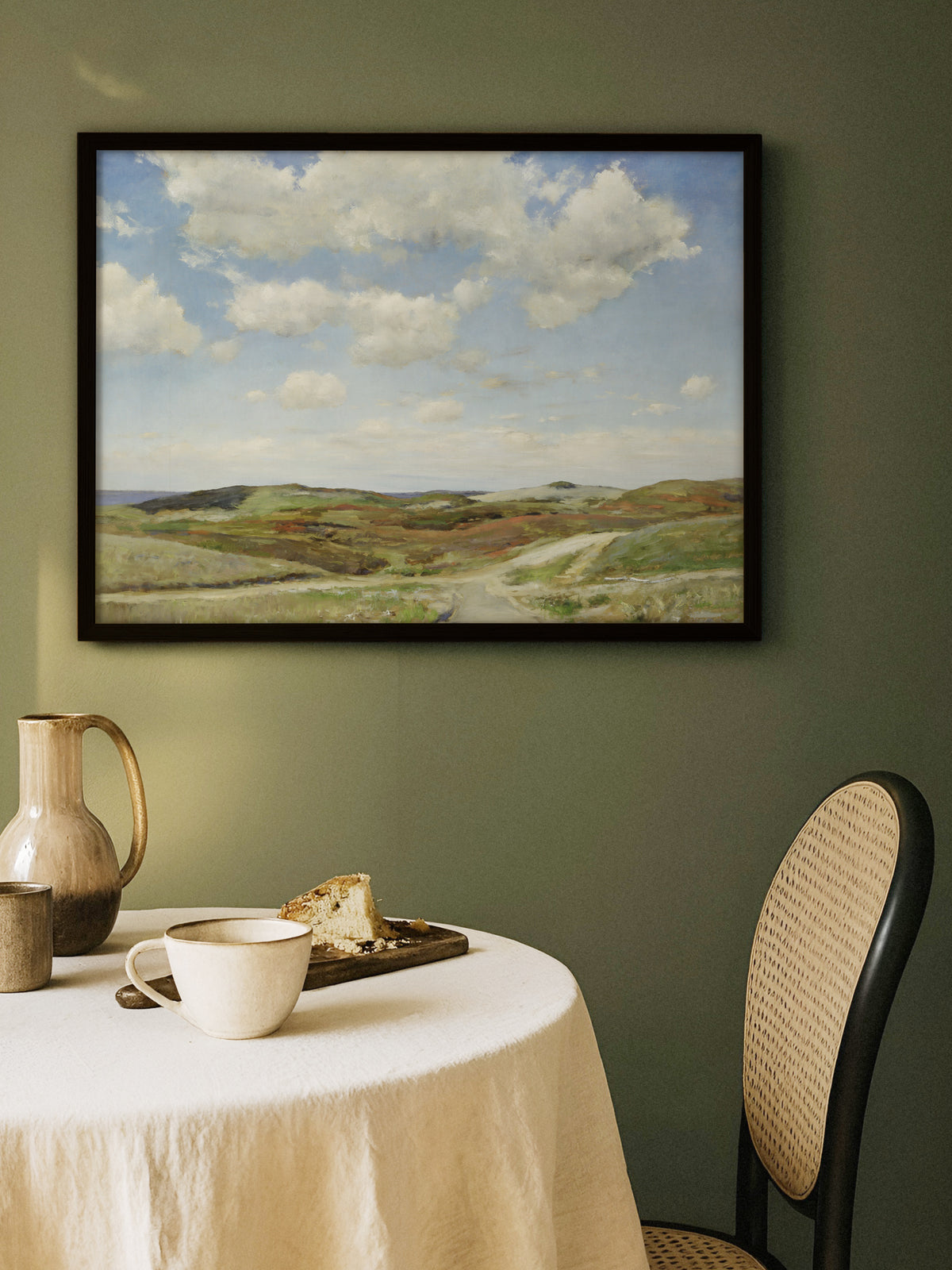 Wandbild mit Dünenlandschaft im schwarzen Rahmen an einer olivgrünen Wand über einem runden Tisch mit Leinentuch, Keramikkrug, Tasse und Kuchen, daneben ein Stuhl mit Wiener Geflecht. Die grünen Hügel passen zur ruhigen Farbe des Esszimmers.