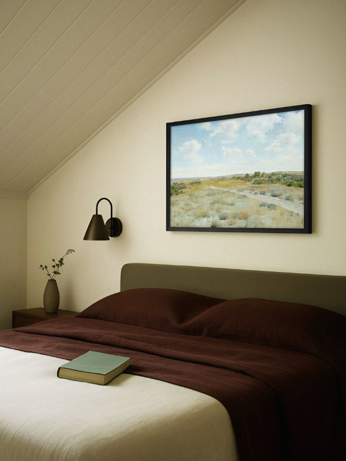 Kunstdruck einer Heidelandschaft im schwarzen Rahmen über einem Bett mit olivgrünem Kopfteil und rotbrauner Bettwäsche unter einer Dachschräge, daneben eine dunkle Wandleuchte und ein Nachttisch mit Vase. Das helle Motiv bringt Himmel in das ruhige Schlafzimmer.