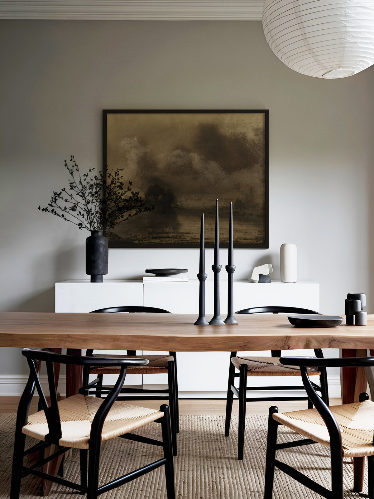 Poster einer Uferlandschaft in Sepia im schwarzen Rahmen über einem weißen Sideboard mit schwarzer Vase, davor ein Holztisch mit schwarzen Kerzenleuchtern und Stühlen mit Papiergeflecht unter einer Papierleuchte. Das dunkle Wandbild setzt einen ruhigen Akzent im Esszimmer.