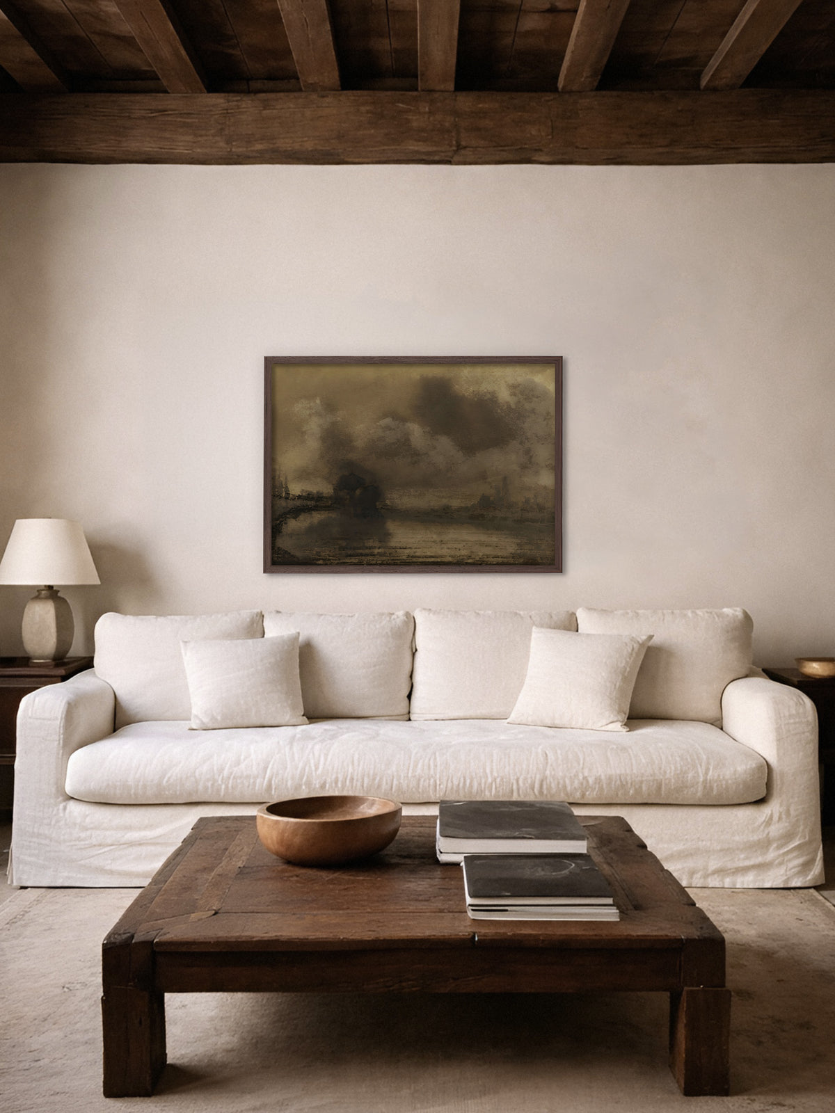 Kunstdruck einer Uferlandschaft in Sepia im dunklen Holzrahmen über einem weißen Leinensofa, davor ein massiver Holztisch mit Holzschale und Büchern, darüber dunkle Holzbalken. Das moody Motiv passt zum rustikalen Wohnzimmer im Wabi Sabi Stil.