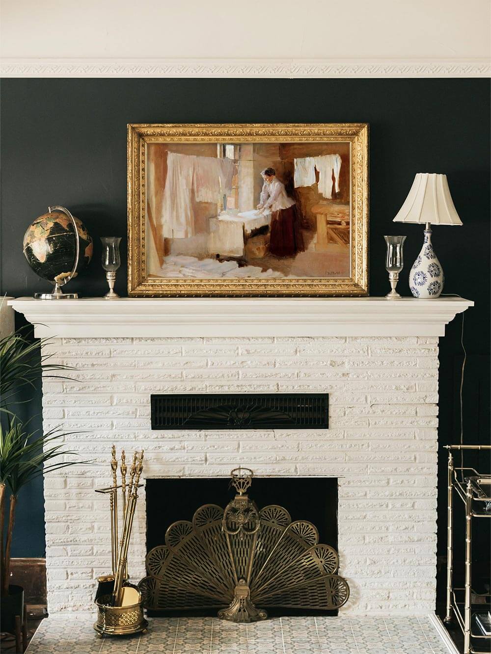 Elegantes Wohnzimmer mit dunkler Wandfarbe und antikem Kaminsims, über dem ein impressionistischer Kunstdruck in einem goldenen Rahmen hängt. Das Gemälde zeigt eine Frau, die Wäsche in einem ländlichen Innenraum ordnet, mit weichen, erdigen Farbtönen.