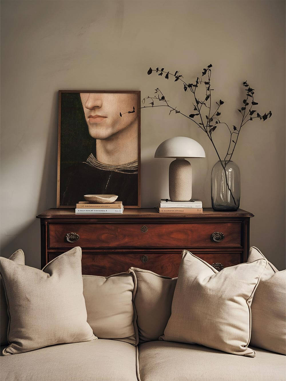 Porträt-Kunstdruck eines jungen Mannes mit feinen Gesichtszügen, dunklem Hintergrund und goldverzierter Kleidung steht auf einer Holzkommode mit Deko aus Glasvase und Büchern. Elegantes Wandbild im Moody Stil für klassische Interieurs.