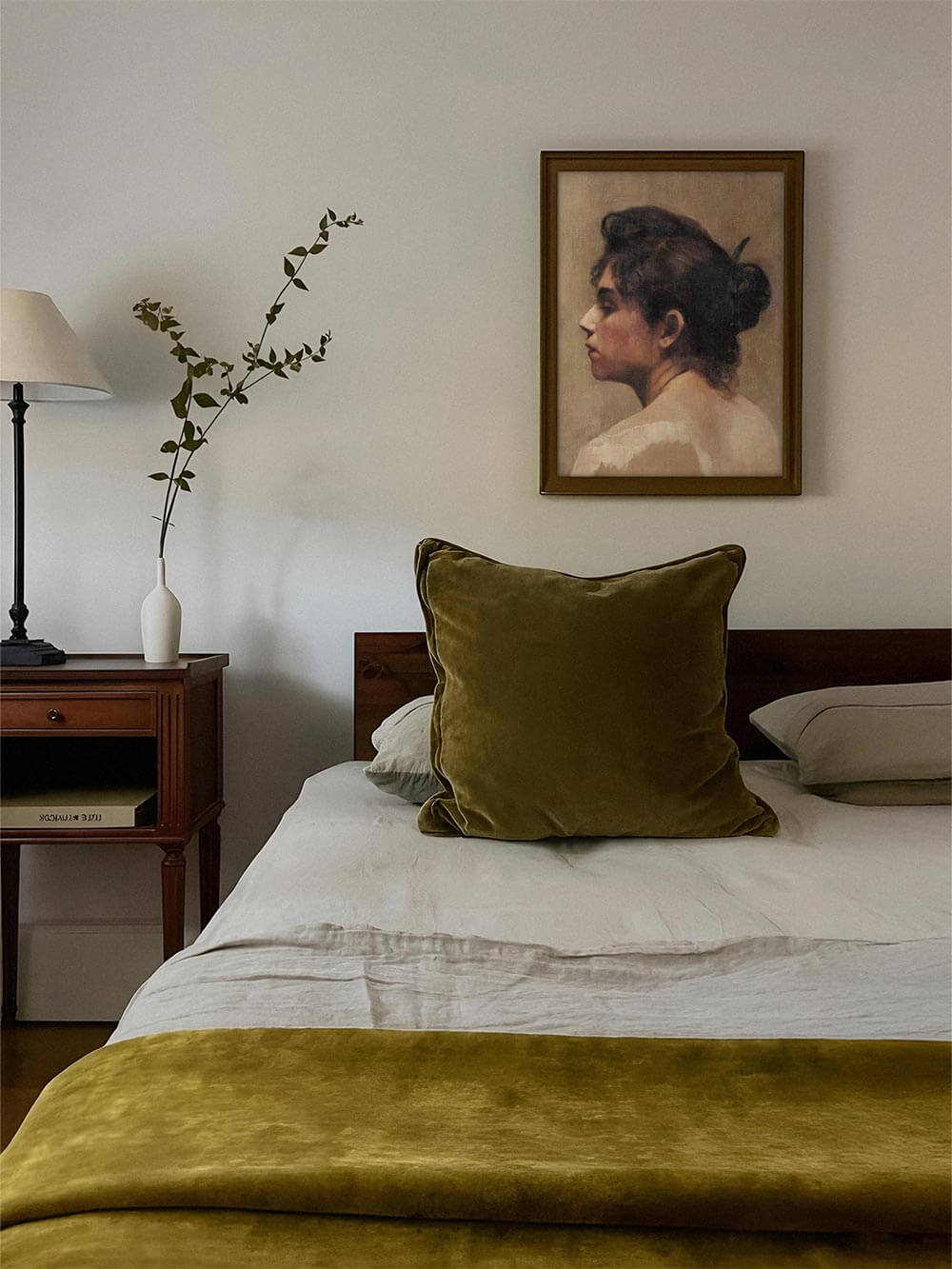 Das Vintage Portrait einer Frau hängt gerahmt über einem Bett mit grünen Akzenten. Der Kunstdruck schafft eine ruhige, nostalgische Stimmung und fügt sich harmonisch ins Schlafzimmer ein.