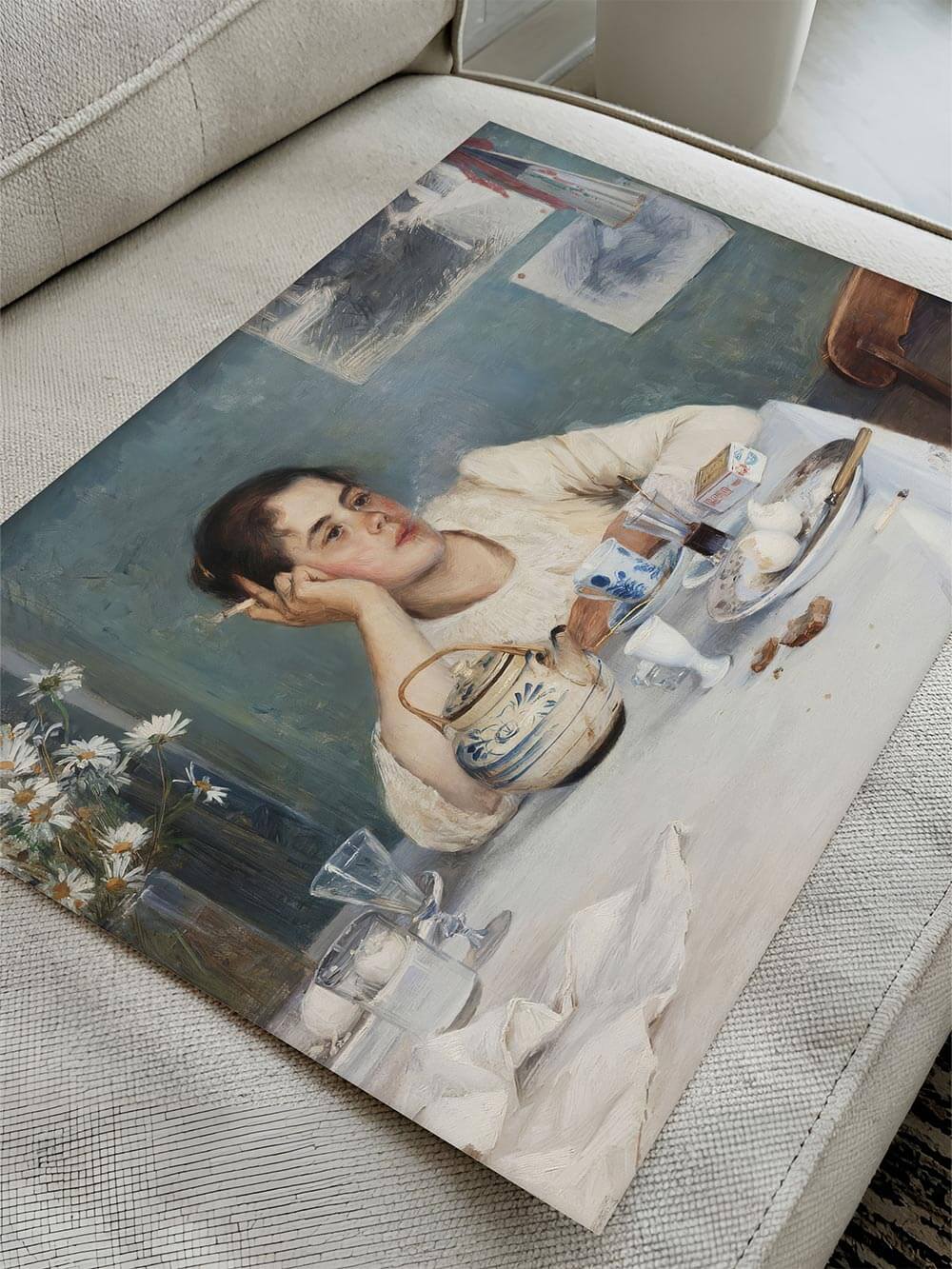 Kunstdruck eines impressionistischen Frauenporträts liegt leicht schräg auf einem hellen Stoffsofa, sichtbar sind weiche Pinselstrukturen, gedeckte Blau- und Cremetöne sowie feine Stillleben-Details, ideal zur Darstellung von Papierqualität und Malstil eines hochwertigen Posters oder Wandbilds.