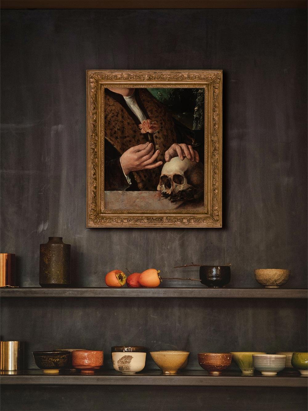 Historischer Kunstdruck mit Schädel und Blume in vergoldetem Rahmen an dunkler Wand mit Keramikvasen. Die Kombination aus Licht und Schatten unterstreicht den klassischen, akademischen Stil.