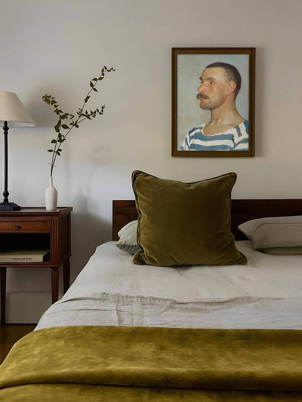 Das Vintage-Porträt eines Mannes hängt gerahmt über einem gemütlichen Bett mit olivgrüner Samtdecke. Das Bild bringt grafische Stärke in das sanft gestaltete Schlafzimmer.
