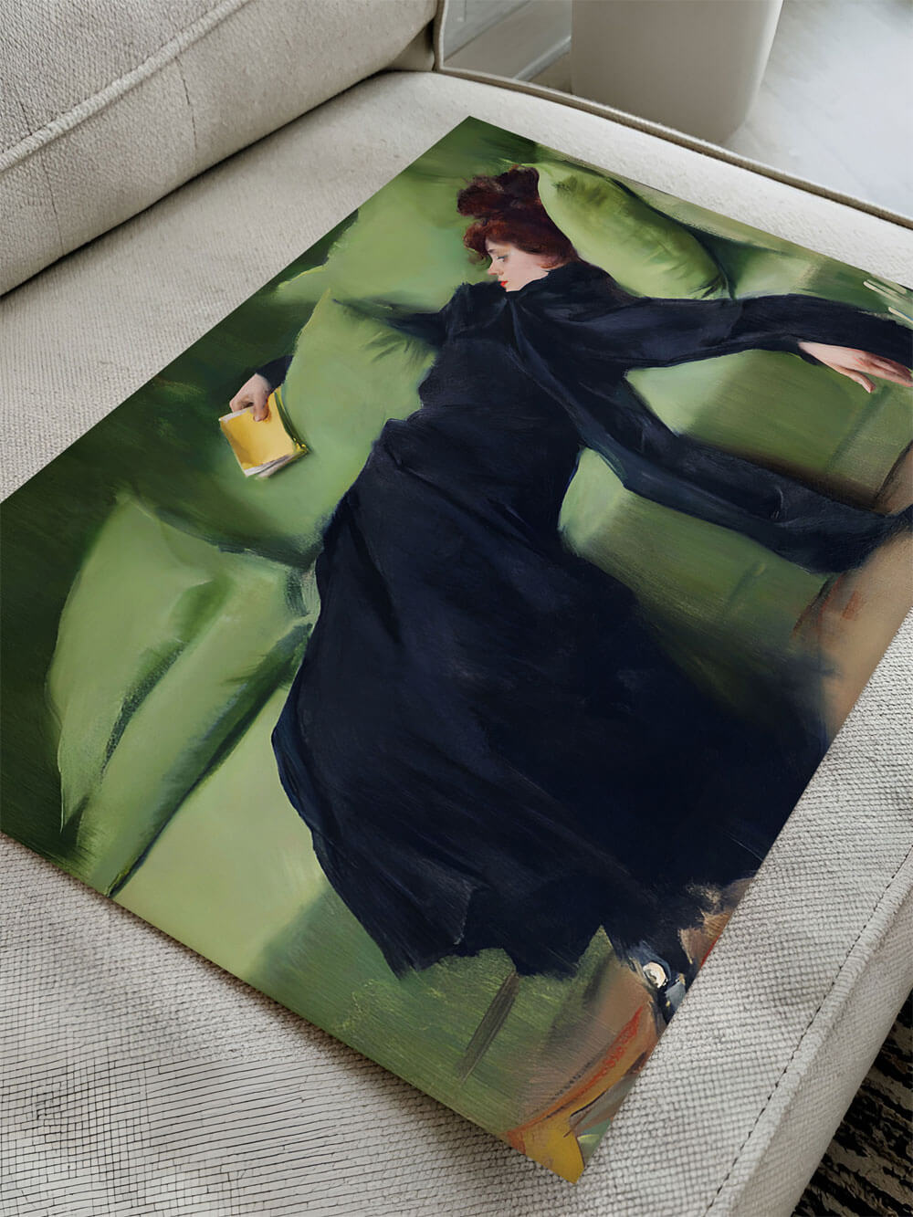 Nahaufnahme eines Kunstdrucks, der eine Frau in schwarzem Kleid zeigt, die auf einem grünen Sofa liegt und ein gelbes Buch hält. Der Druck liegt auf einem hellen, strukturierten Sofa, das Detail und die Farbtiefe des Bildes hervorhebt.