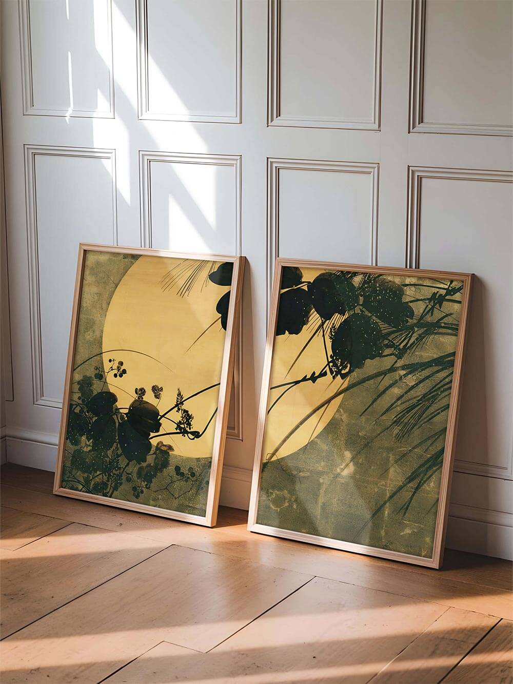 Zwei gerahmte Kunstdrucke lehnen an einer hellen Wand im Sonnenlicht. Die Kombination aus Holzrahmen, goldgelben Tönen und zarten Schatten vermittelt Balance und Eleganz im modernen Wabi-Sabi-Interior.